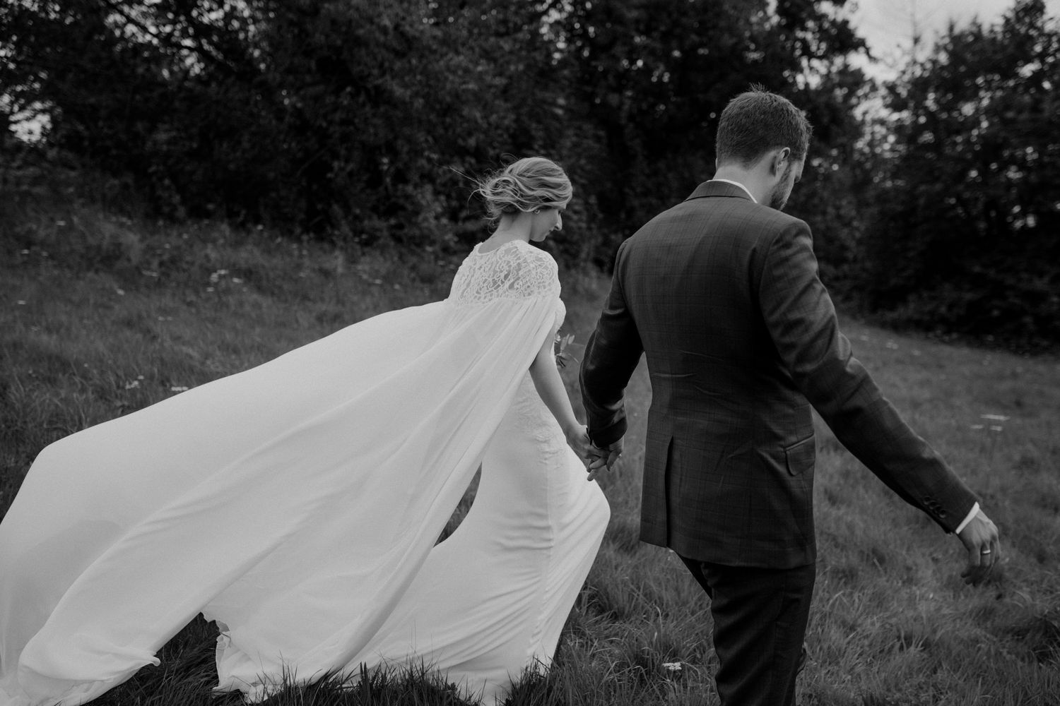 Black and white photo of bride with flowing cape veil and groom walking through green countryside.