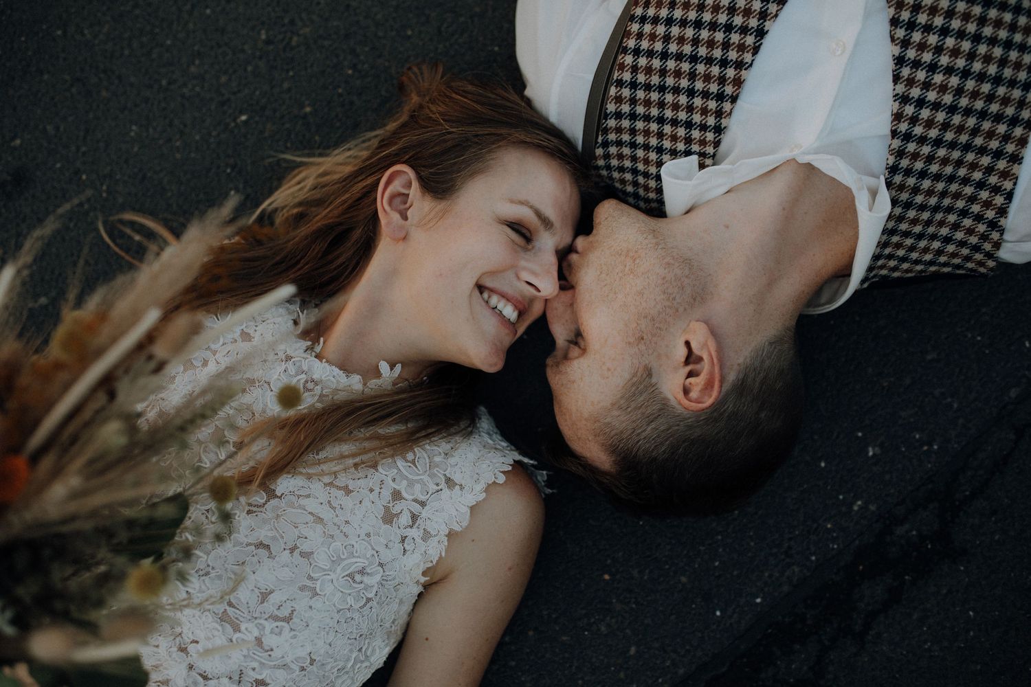 Overhead view of smiling bride and groom lying head to head on ground with dried floral arrangement.