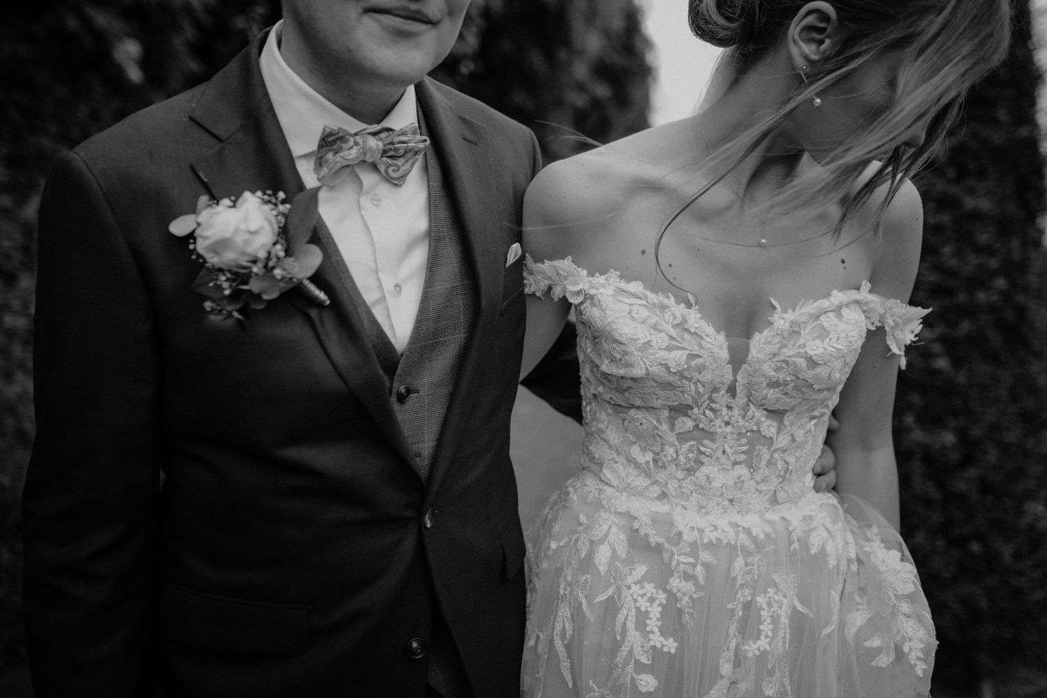Black and white close-up of bride in floral lace gown and groom in suit with bow tie boutonniere.