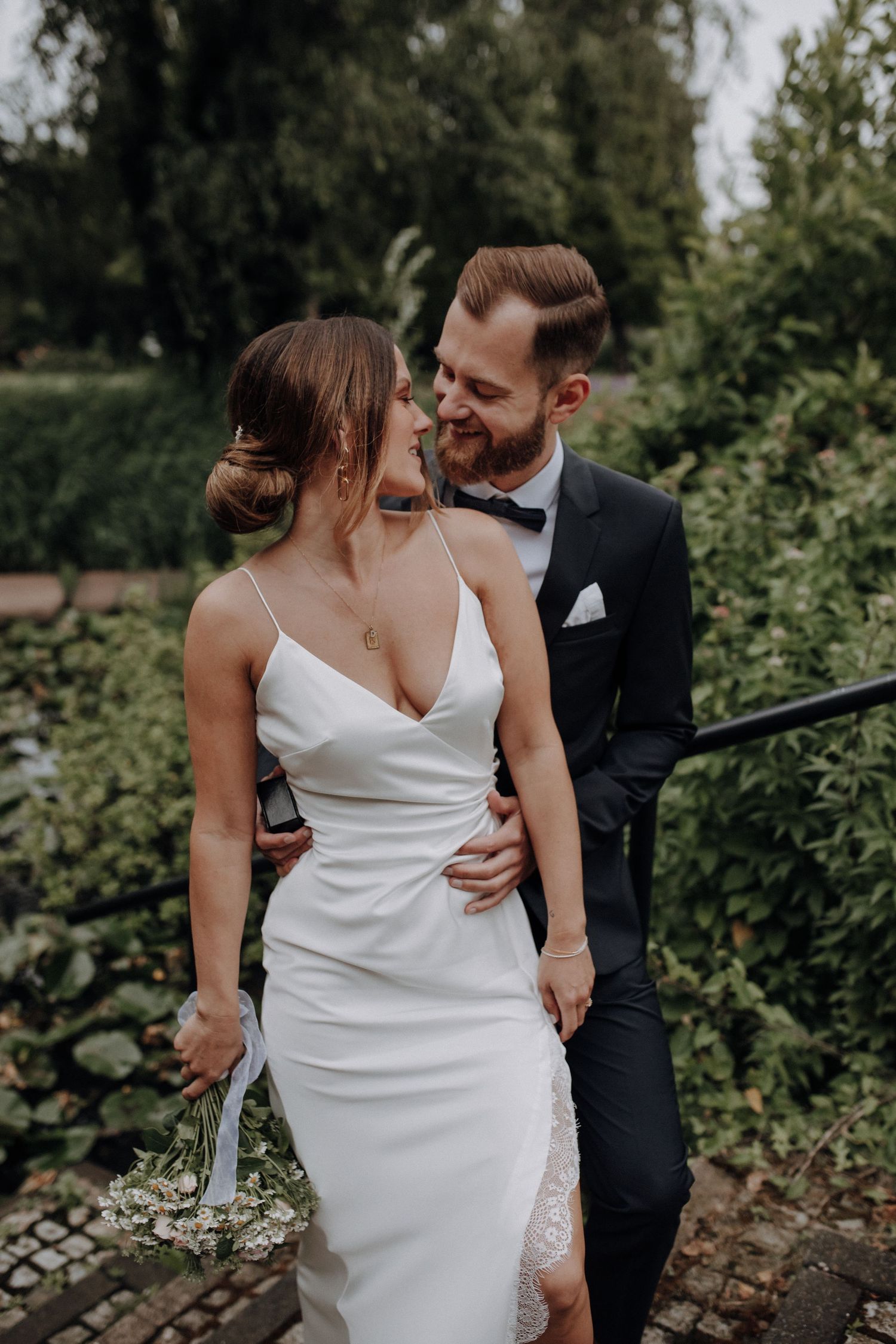 Stylish couple in wedding attire smiling intimately surrounded by lush green garden foliage.