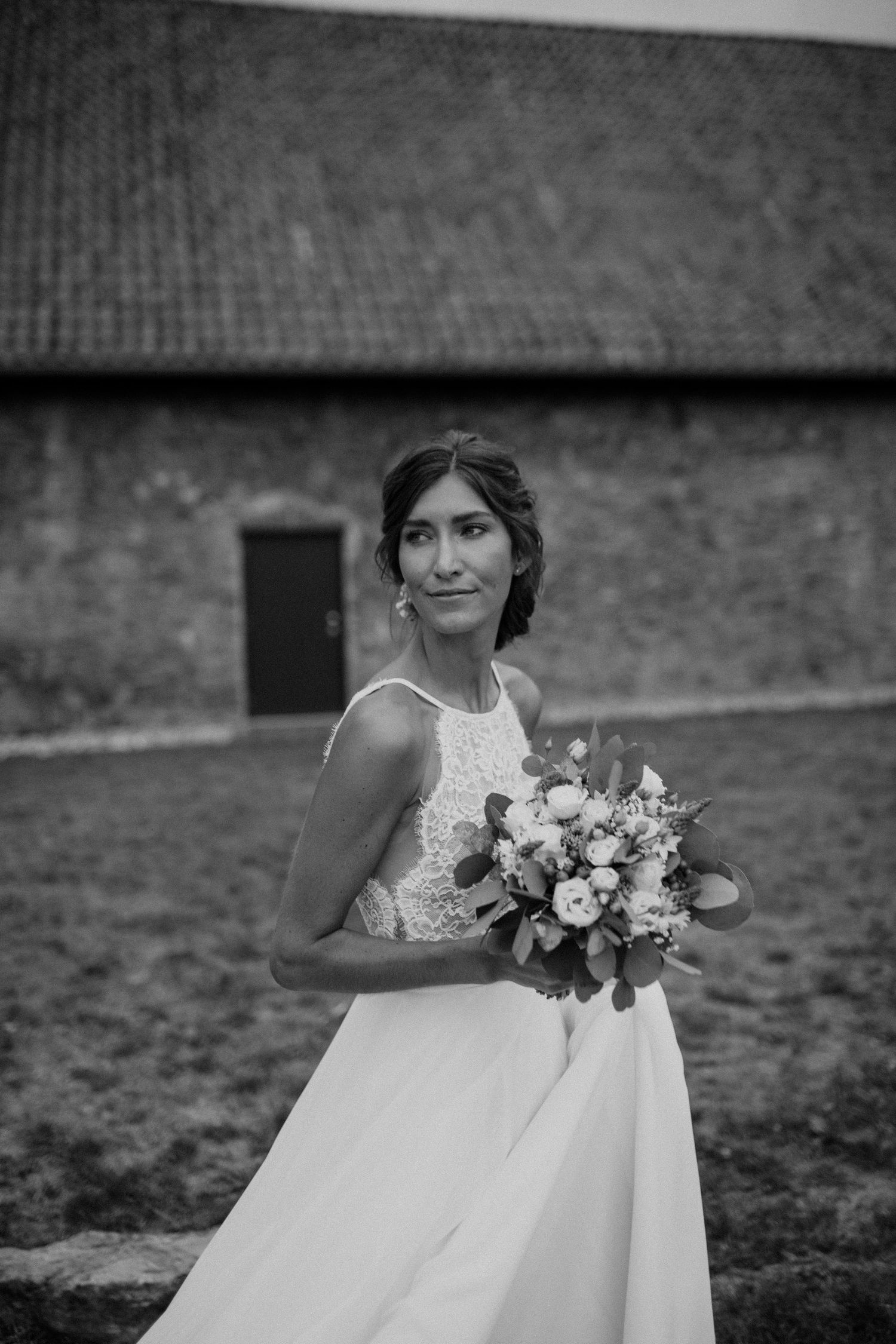 Black and white portrait of bride holding floral bouquet standing before a rustic stone barn.