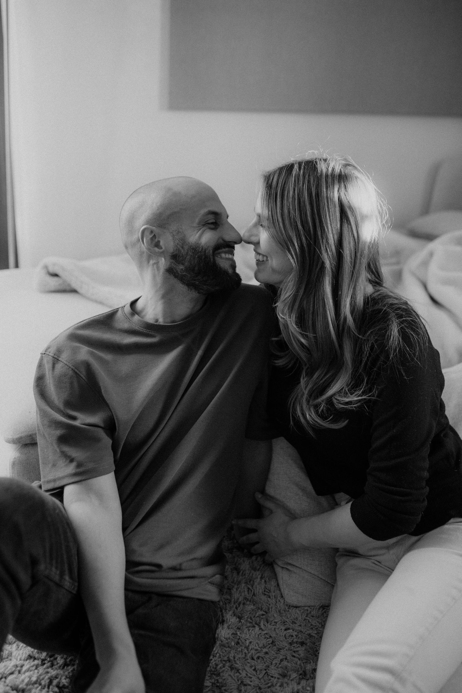 Black and white photo of a loving couple sharing a playful nose-to-nose moment near a bed.