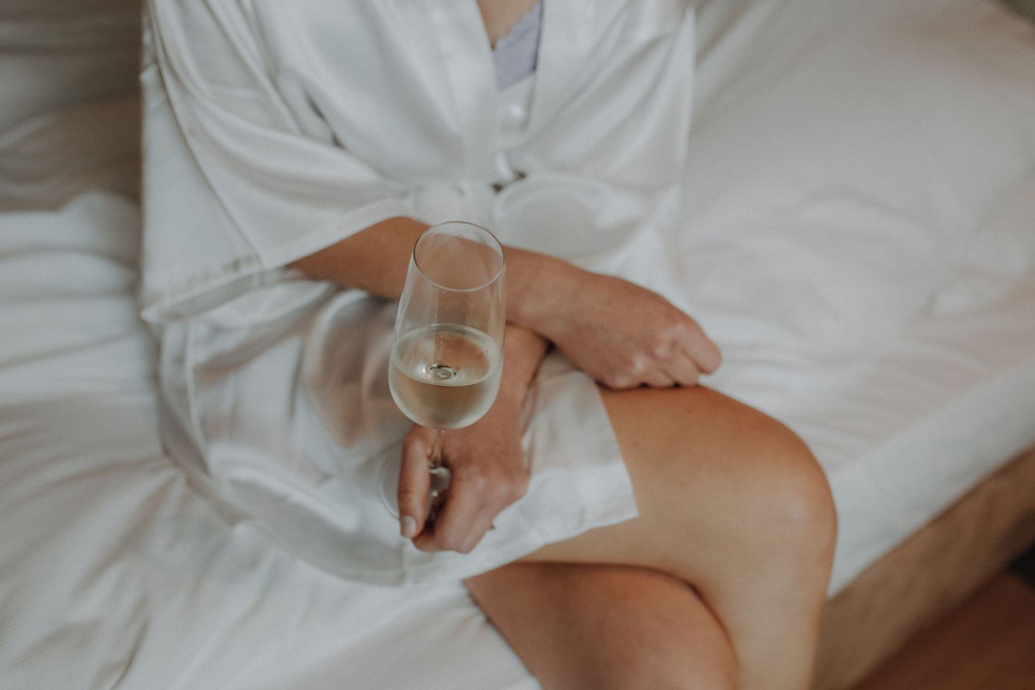 Woman in white silk robe sitting on bed holding a glass of champagne on her wedding day.