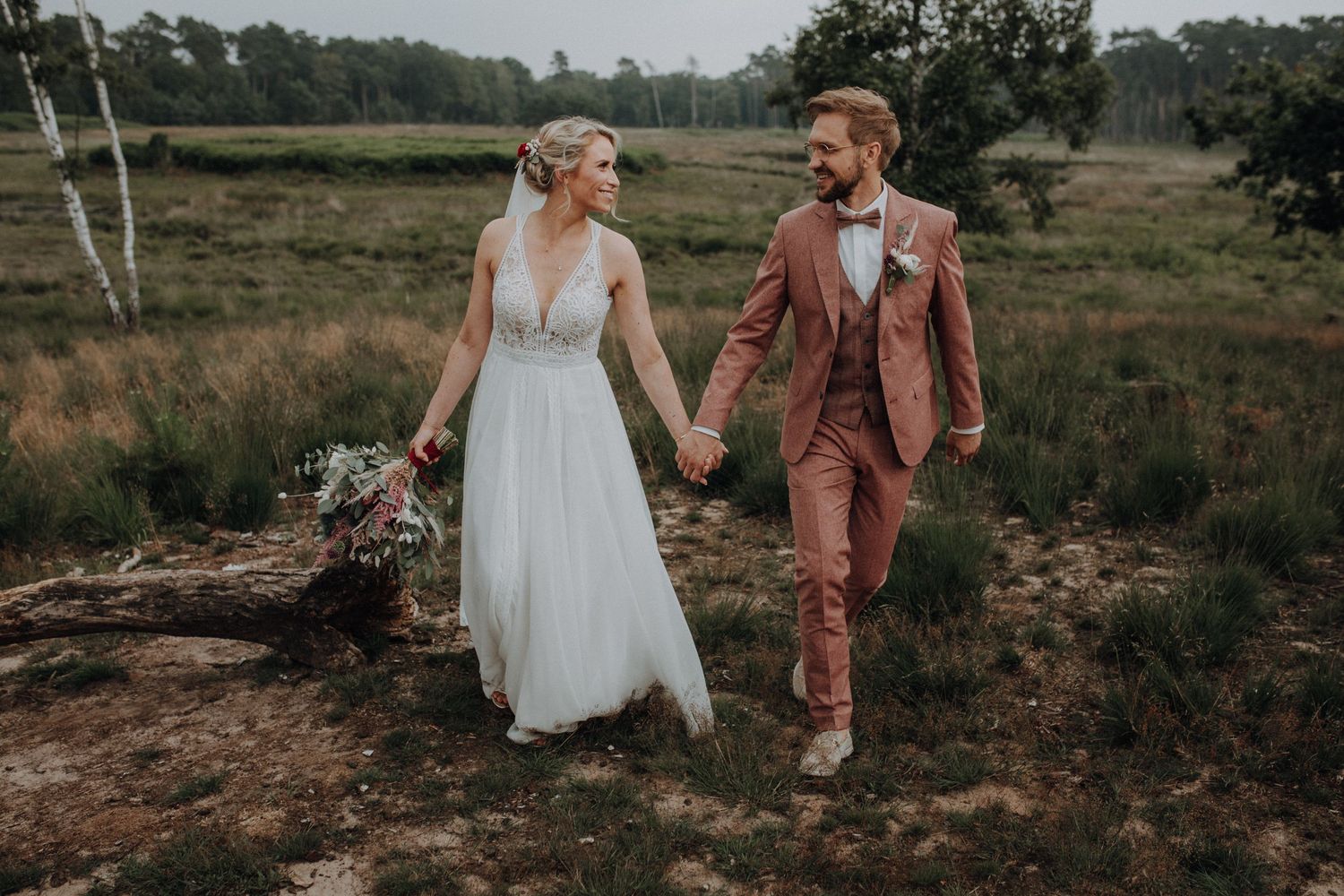 Happy bride and groom in pink suit strolling hand-in-hand through a lush green meadow.