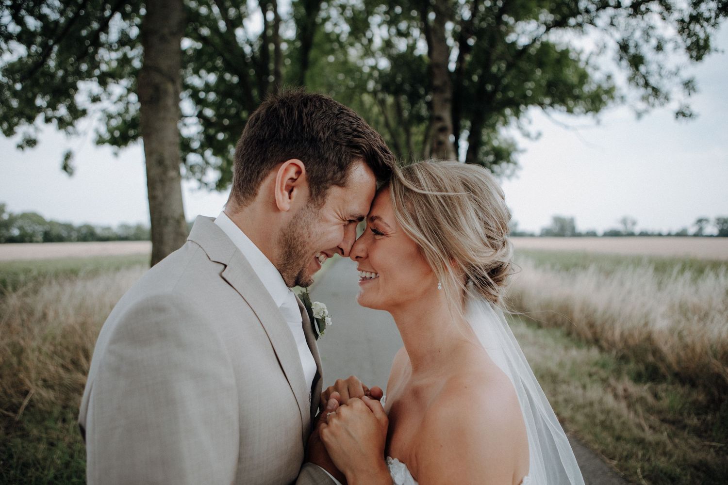 Joyful bride and groom in neutral tones touching foreheads tenderly beneath tall trees in field.
