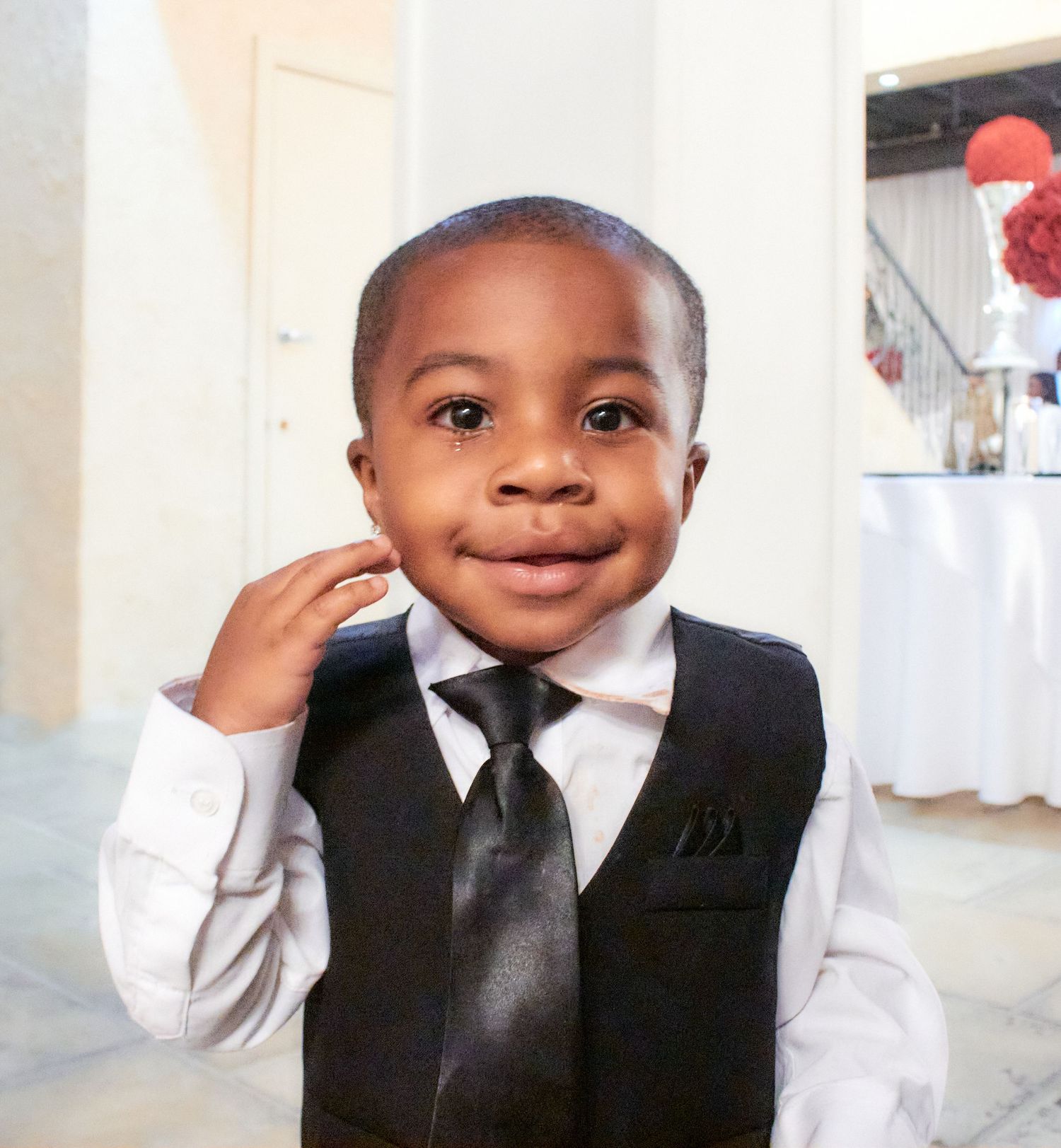 Young child in formal black suit vest and tie smiling at camera against light background.