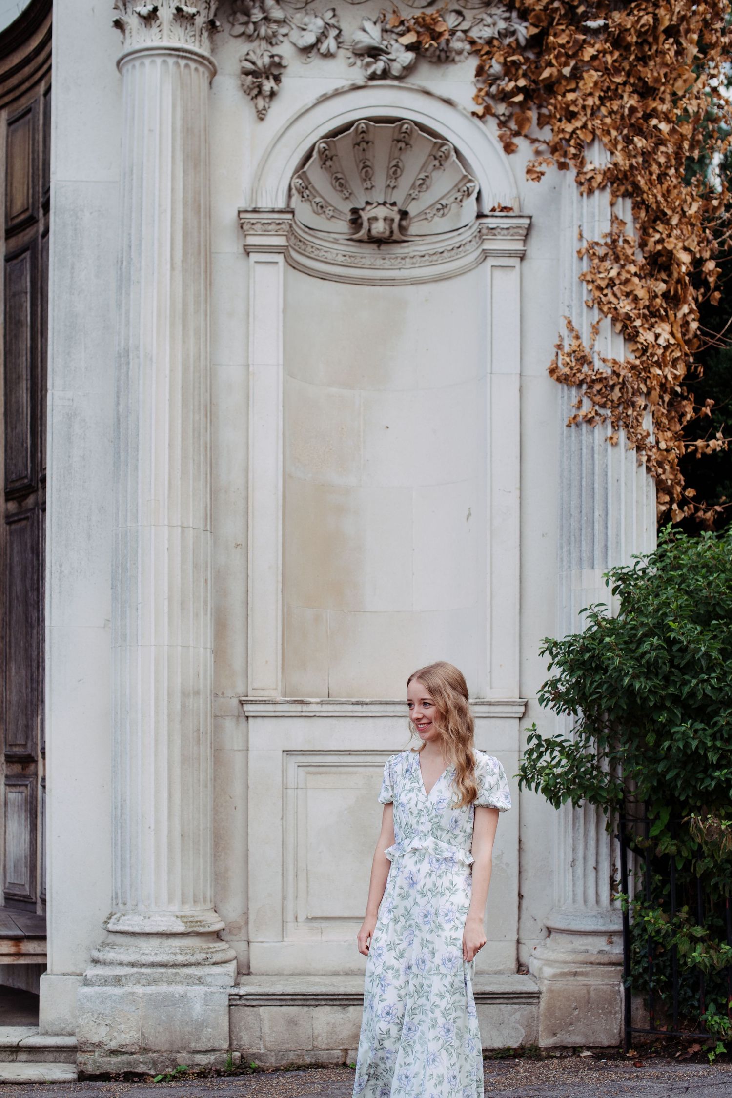 A woman in a floral dress stands elegantly against white classical architecture with columns and autumn foliage in the background.