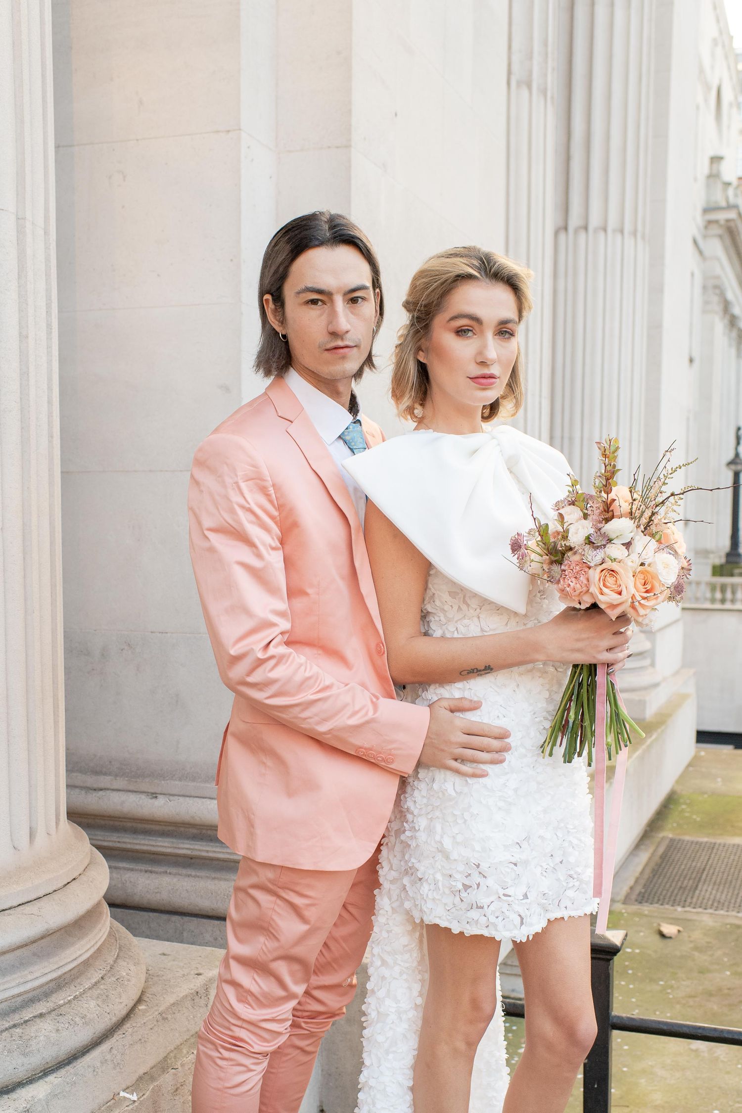 A couple poses for a wedding photo outdoors, with the person in a pink suit and their partner wearing a white dress.