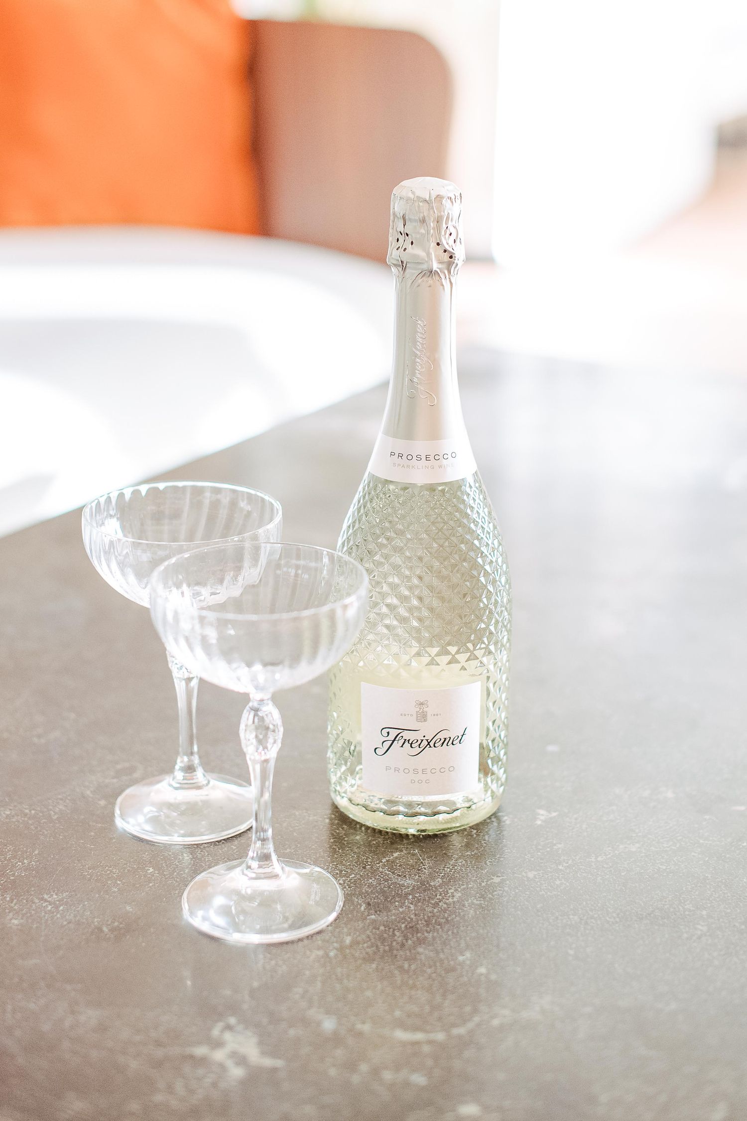 Two crystal champagne coupes and a decorative white glass bottle sit on a textured grey surface in soft lighting.