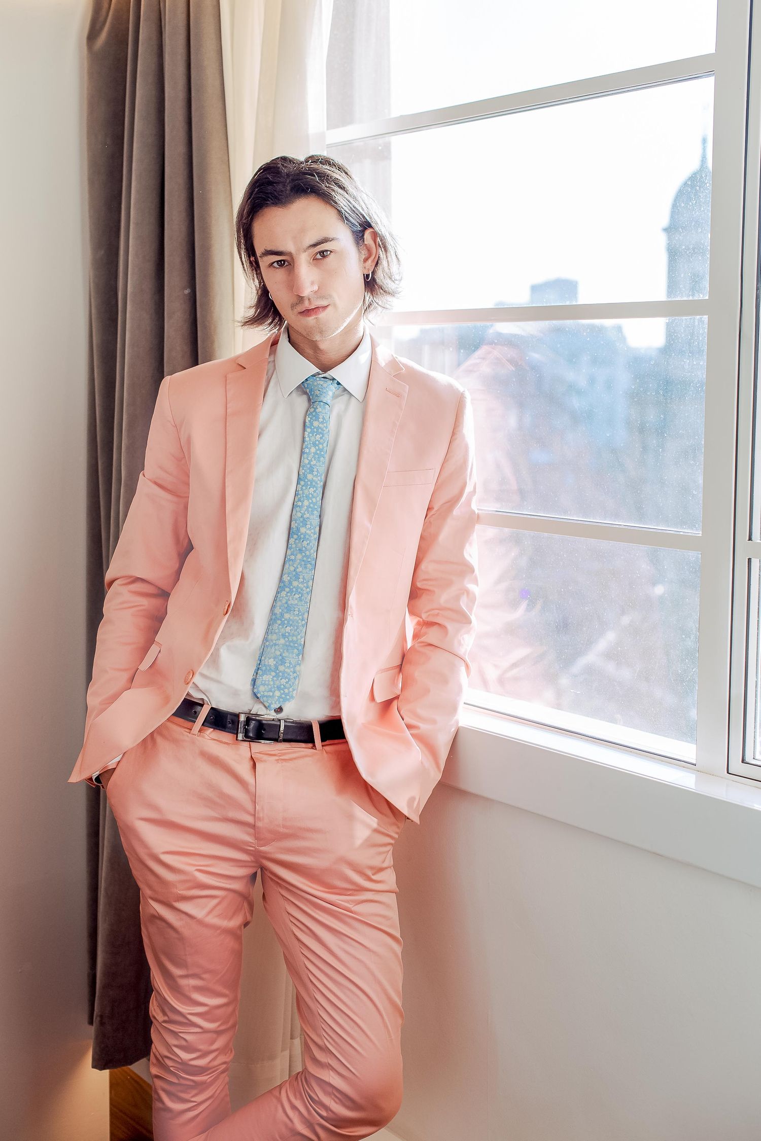 Person in stylish salmon pink suit with light blue patterned tie poses near window in natural light.