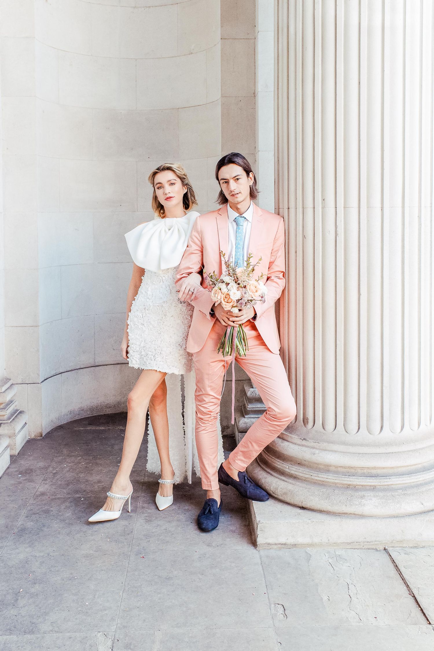 Modern fashion editorial of a wedding couple posing in a white dress and salmon pink suit by classic columns at the Maryelbone Town Hall.