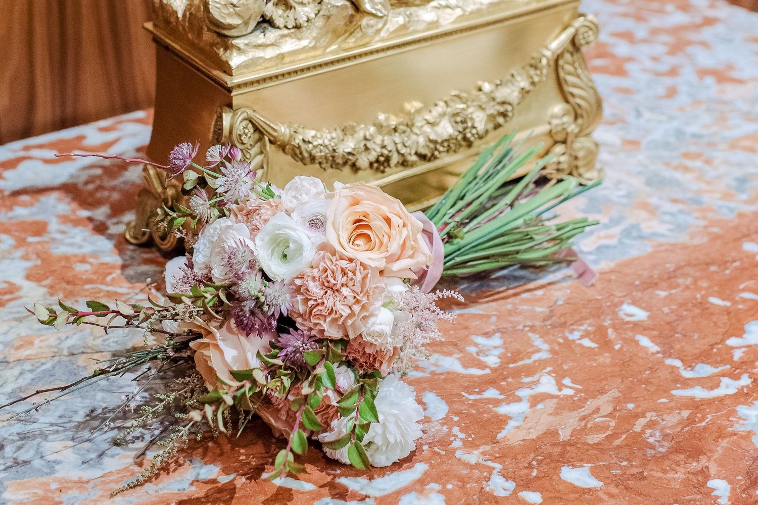 An elegant wedding bouquet with peach roses and mixed flowers rests beside a decorative gold antique box on marble surface.