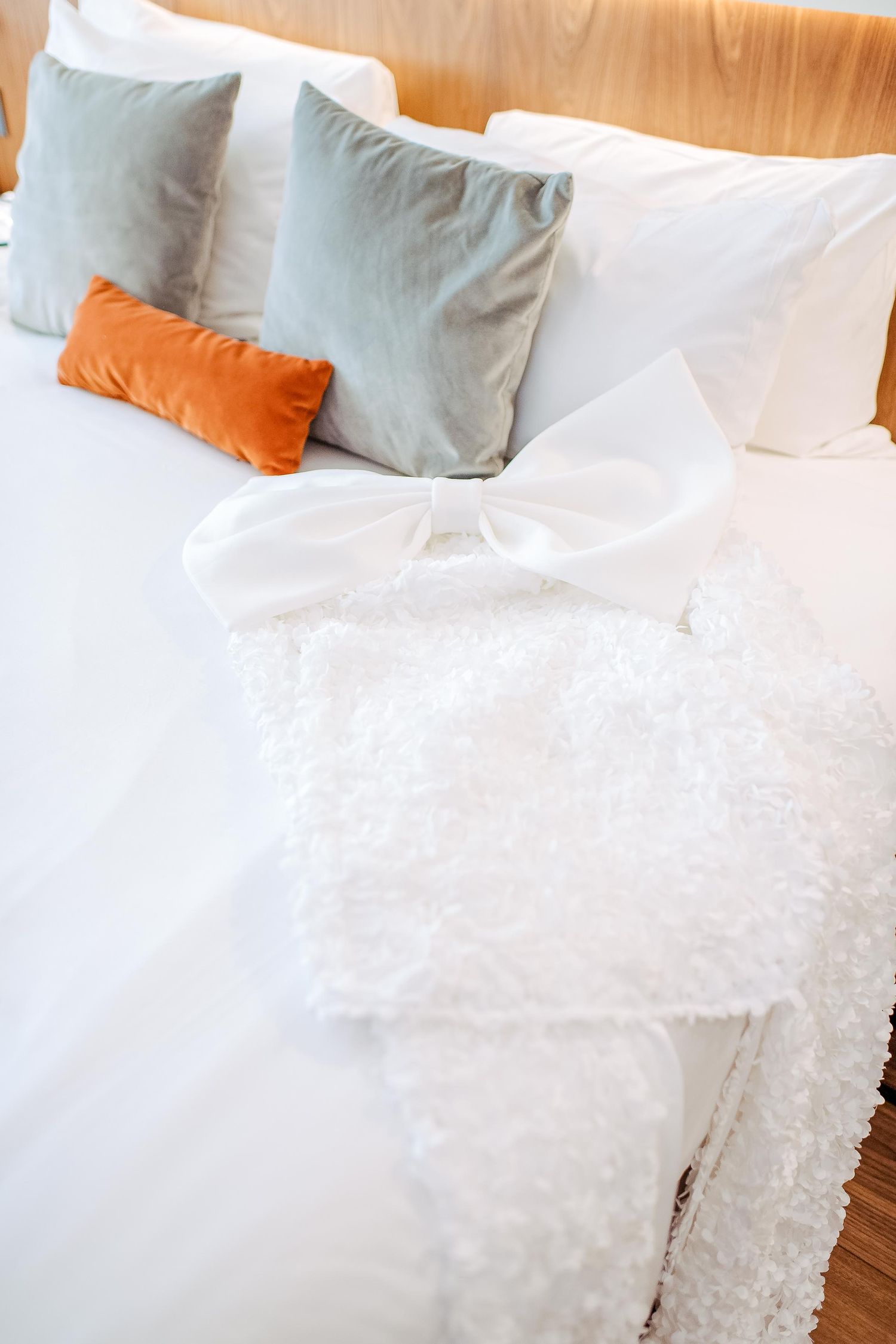 A white bed with grey and orange accent pillows features a delicate lace wedding dress laid out on top.