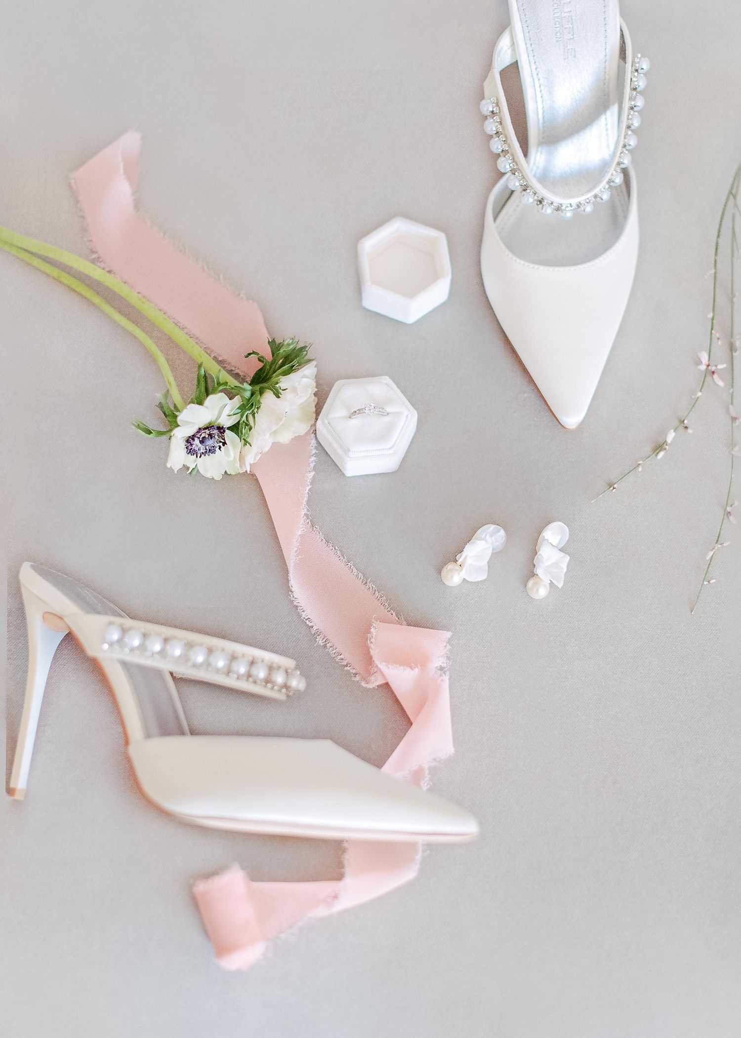 Elegant white pearl-studded wedding heels with pink ribbons and accessories on a grey surface.