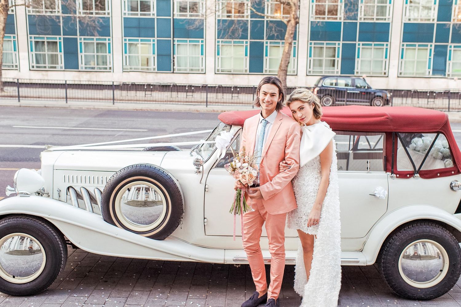 A classic white vintage luxury car parked on a London street with a couple in pastel-colored outfits posing beside it on their wedding day.