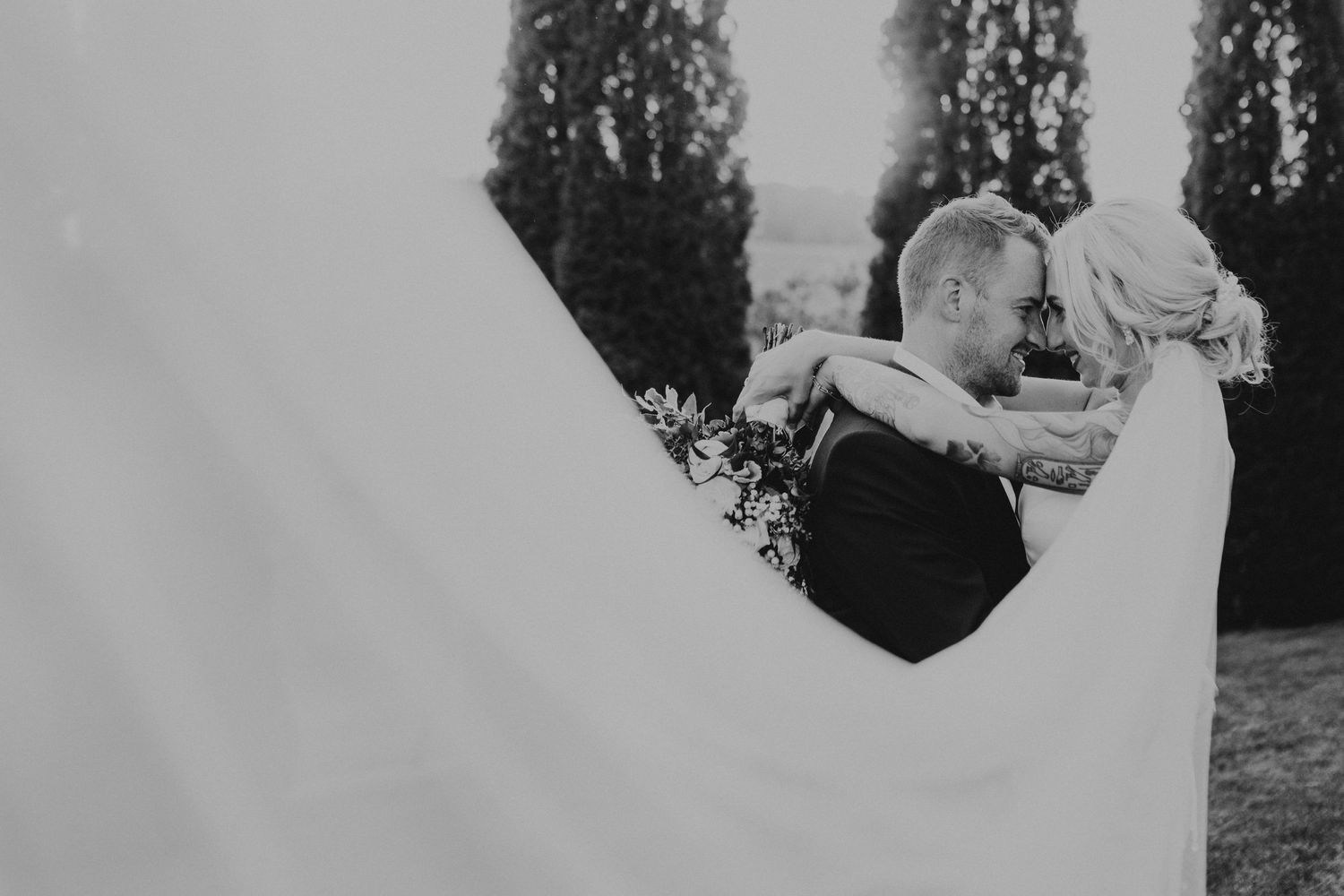 A tender wedding moment captured among cypress trees in black and white.