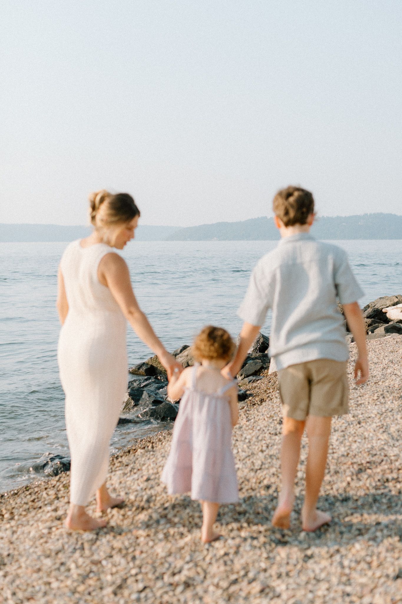 Family of three walking hand-in-hand on a rocky beach, parents in white and beige, young girl in pink dress.