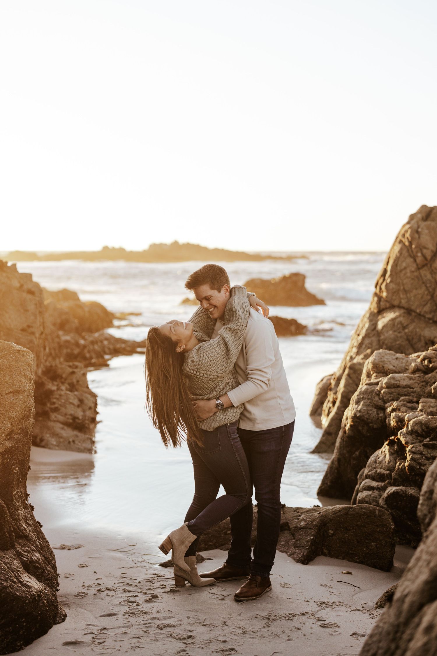 couple embracing and laughing on a beach with rocks surrounding them and golden light peeking through