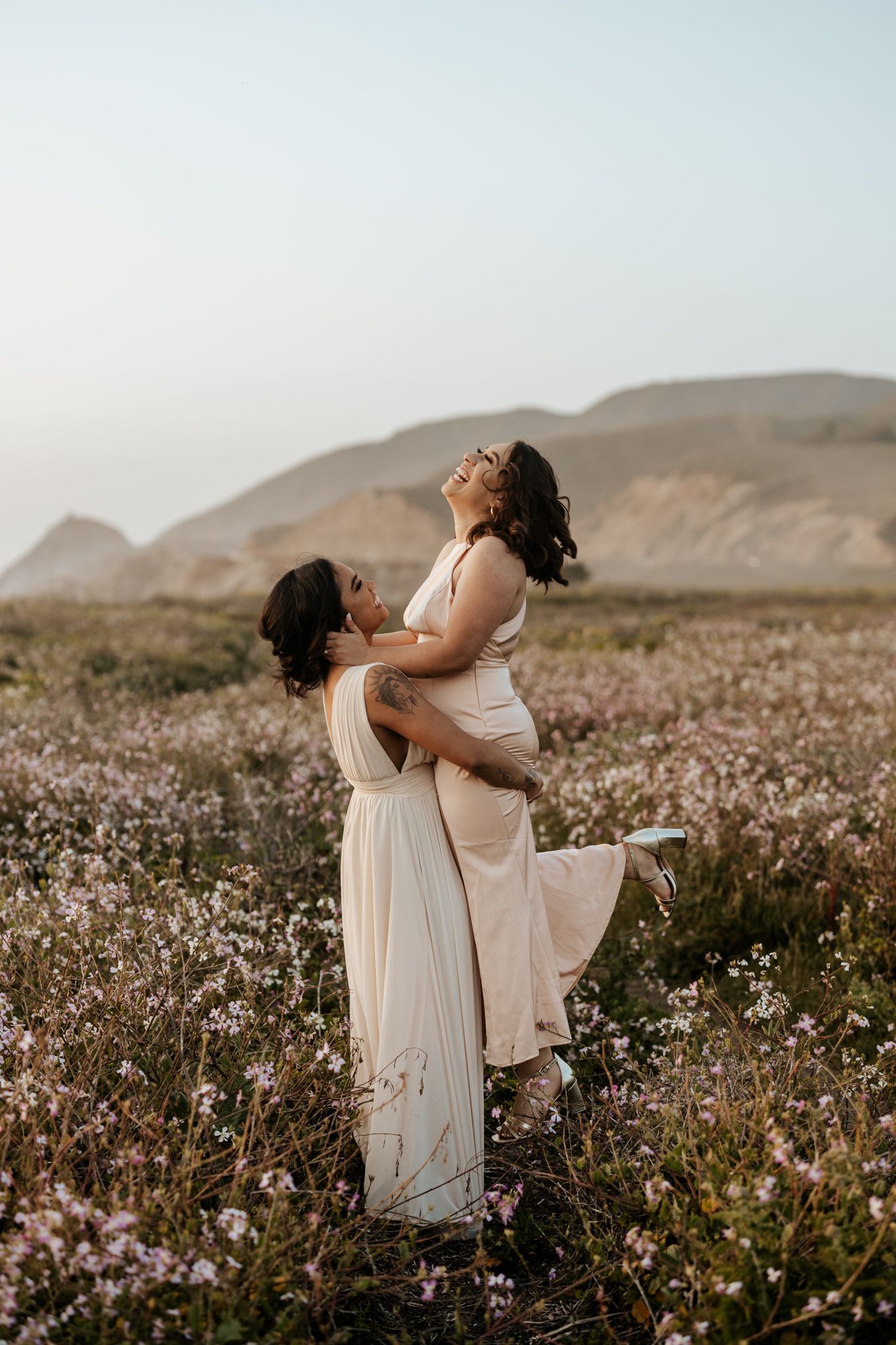 two women in a field of flowers on a cliff with a mountain behind them, lifting each other and laughing