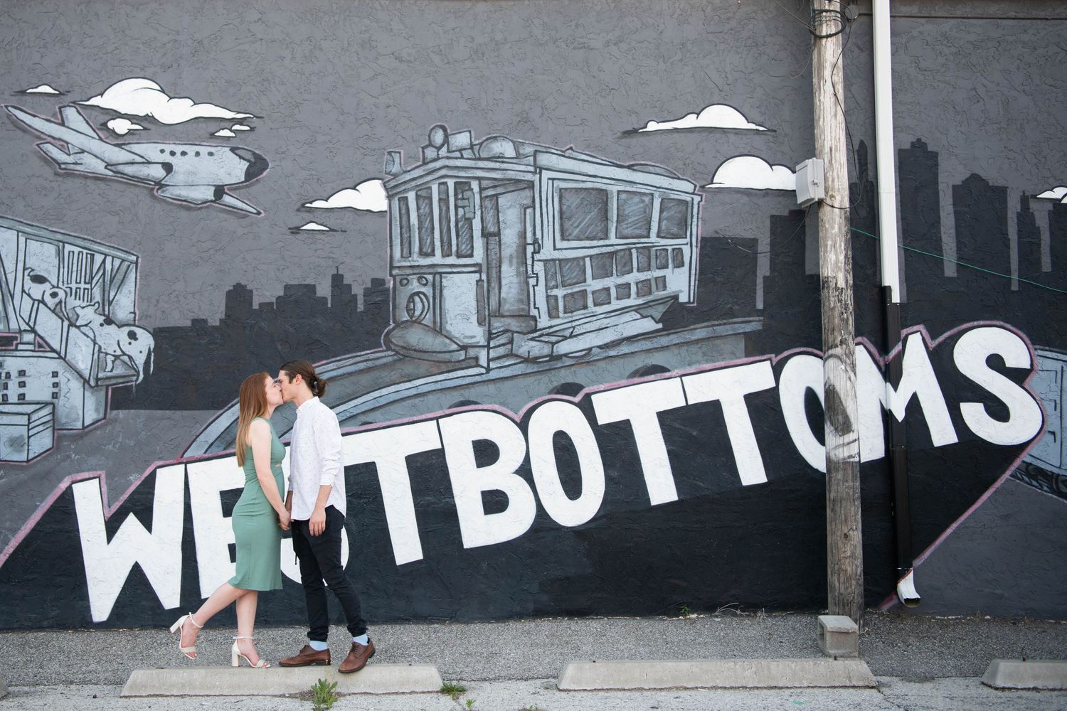 A couple stands before a urban mural reading 'Westbottoms' in black and white.