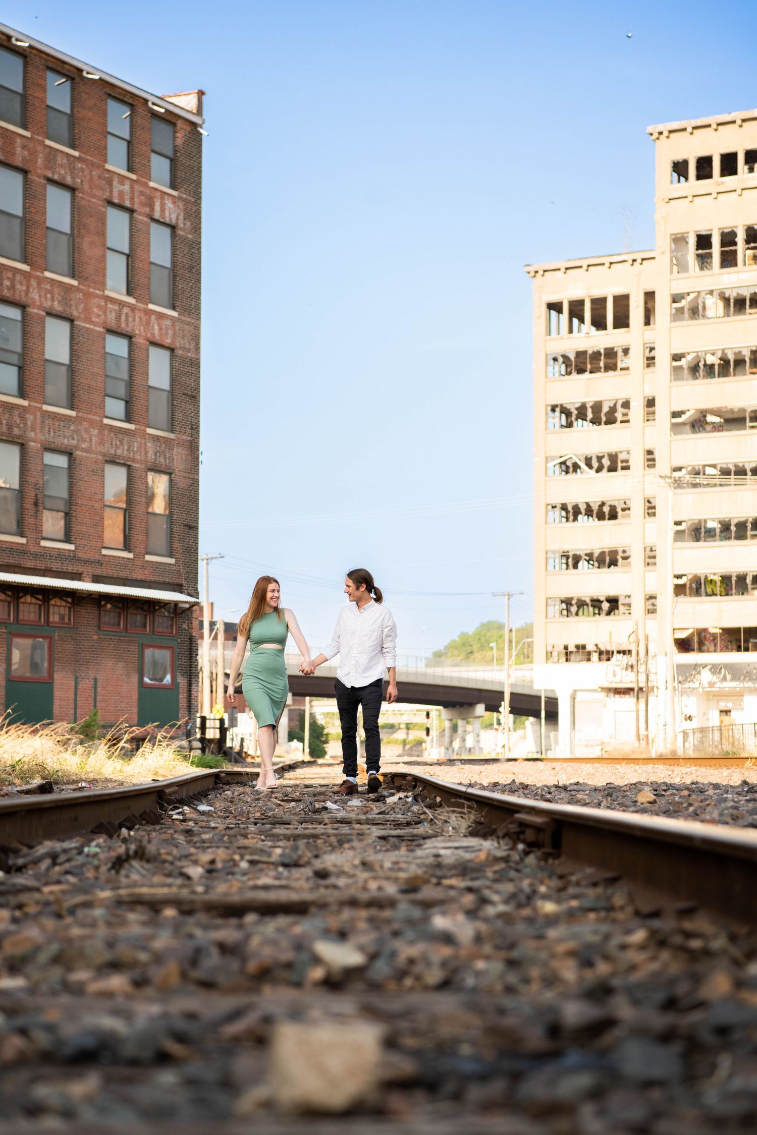 Couple walking along railroad tracks between brick buildings in an urban setting.