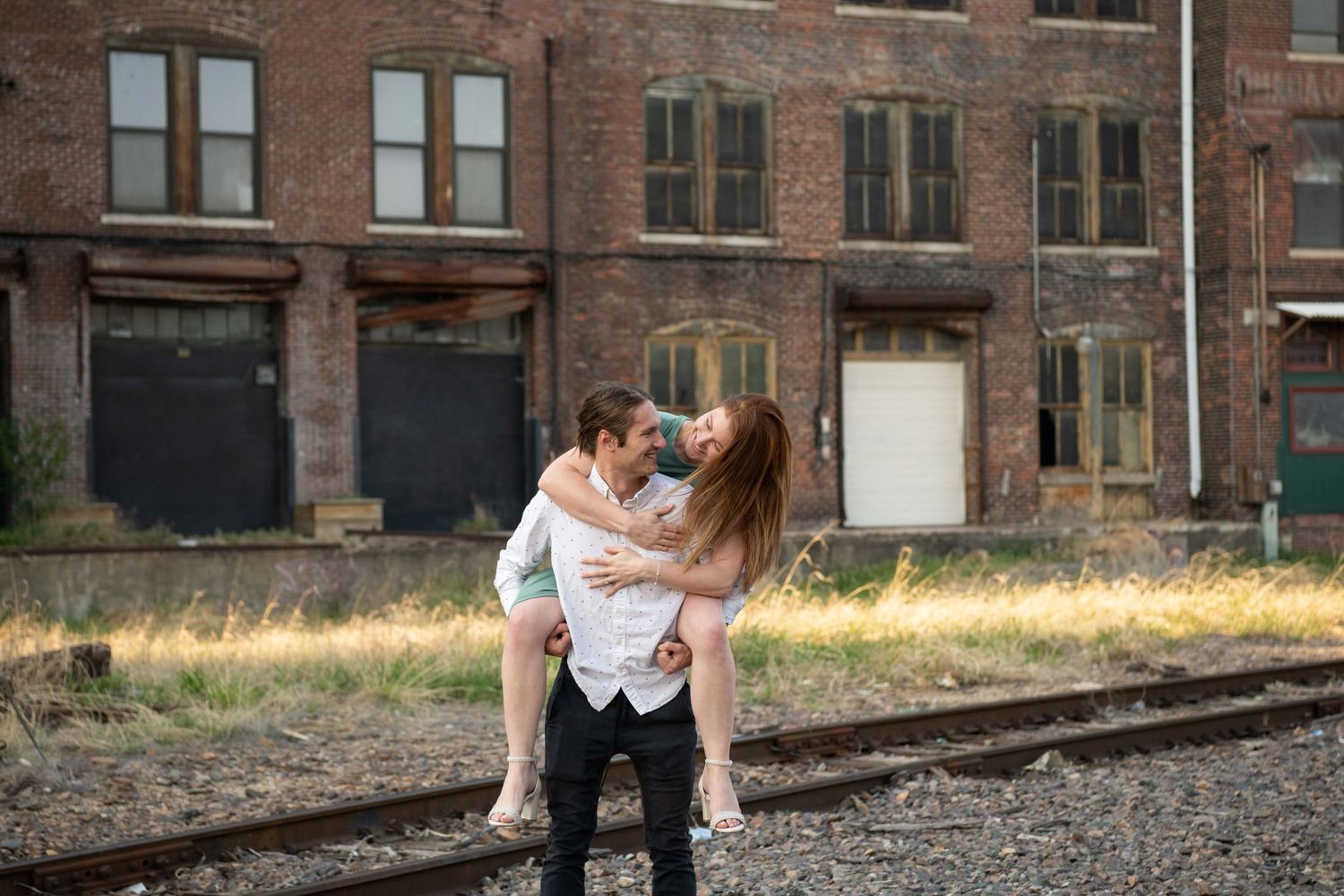 Playful moment between couple on railroad tracks with brick building backdrop.