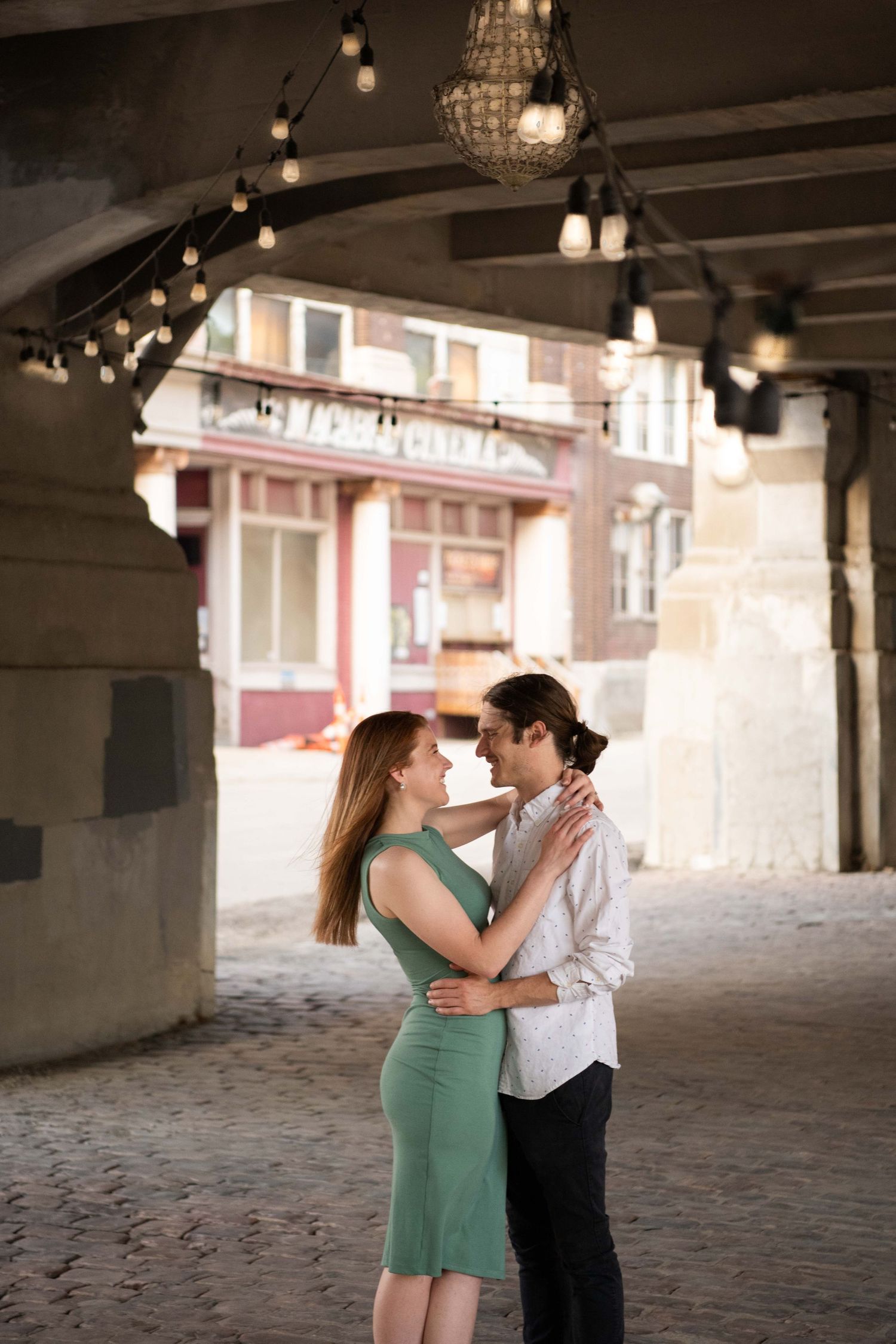 A romantic couple embraces under string lights in an arched passageway at dusk.