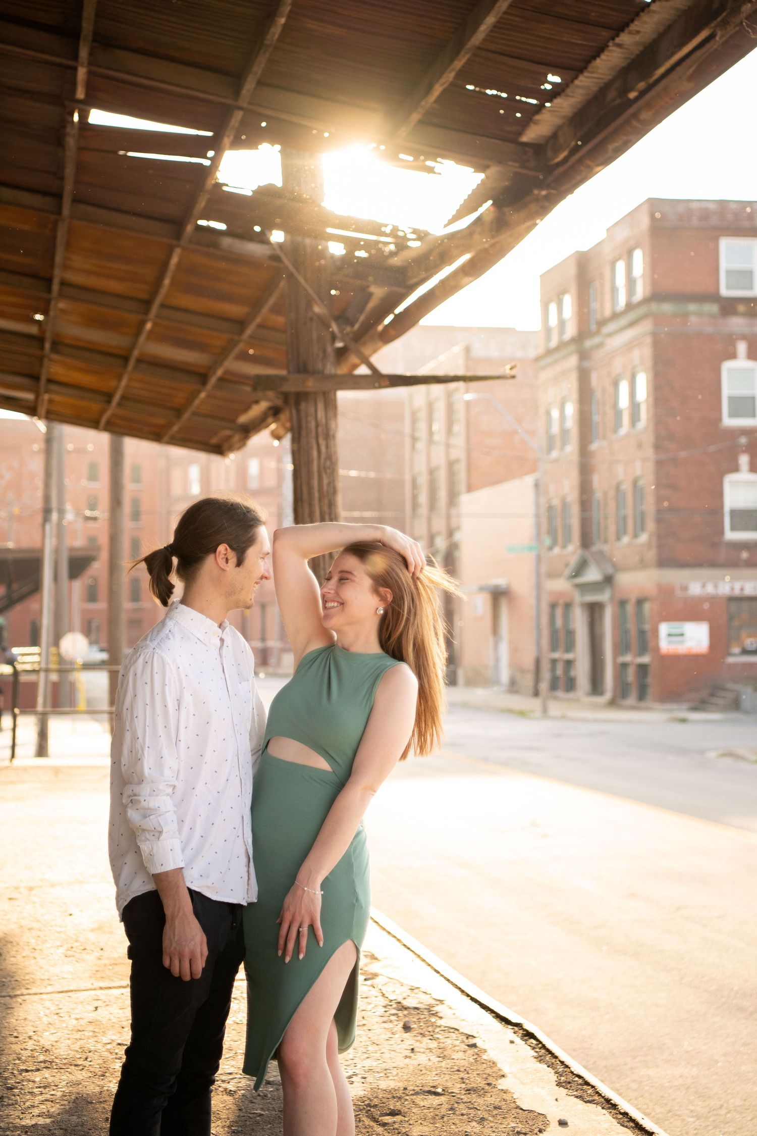 A tender moment between a couple standing beneath a wooden overhang with sunlit brick buildings behind them.