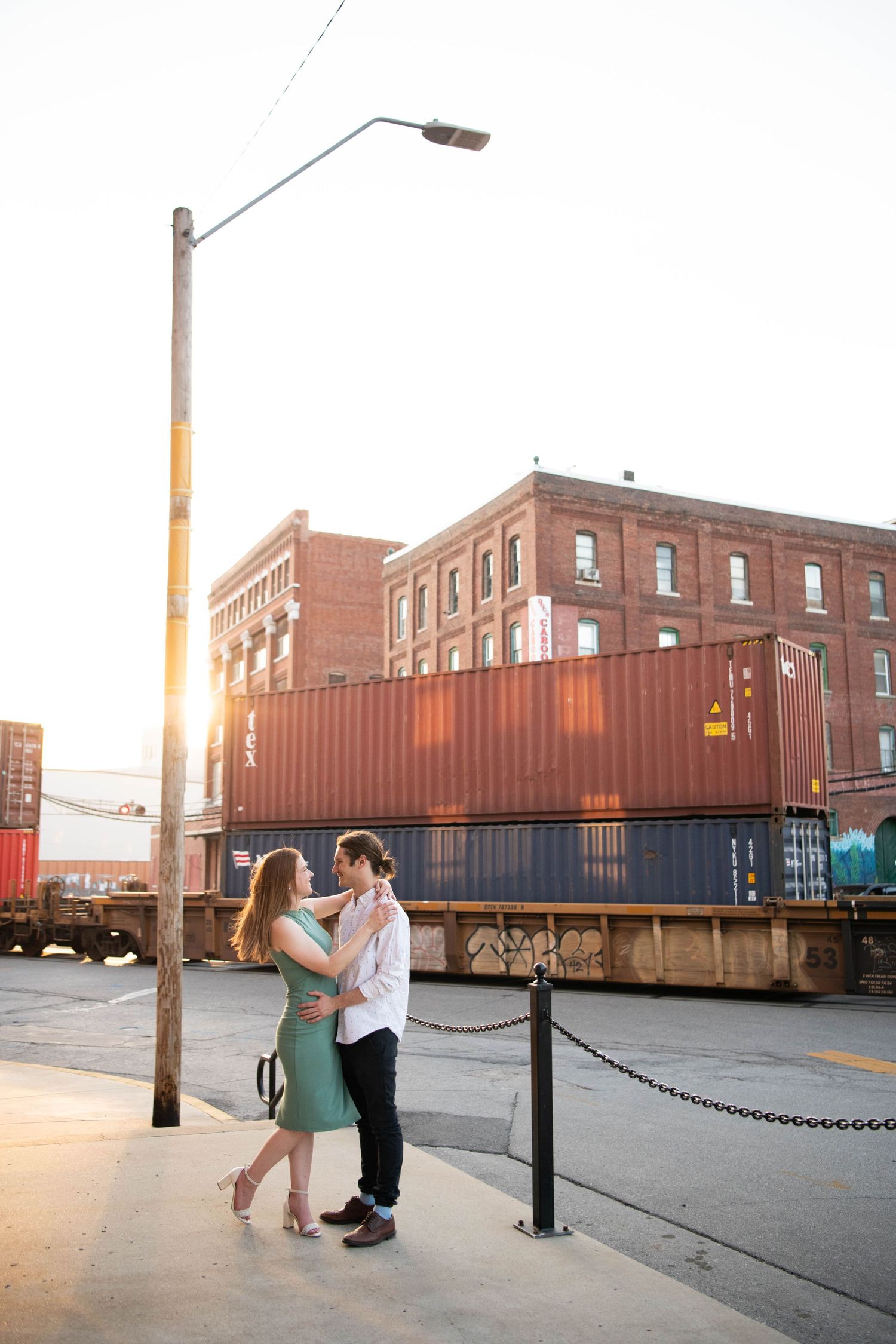 A romantic couple poses near stacked shipping containers at sunset in an urban setting.