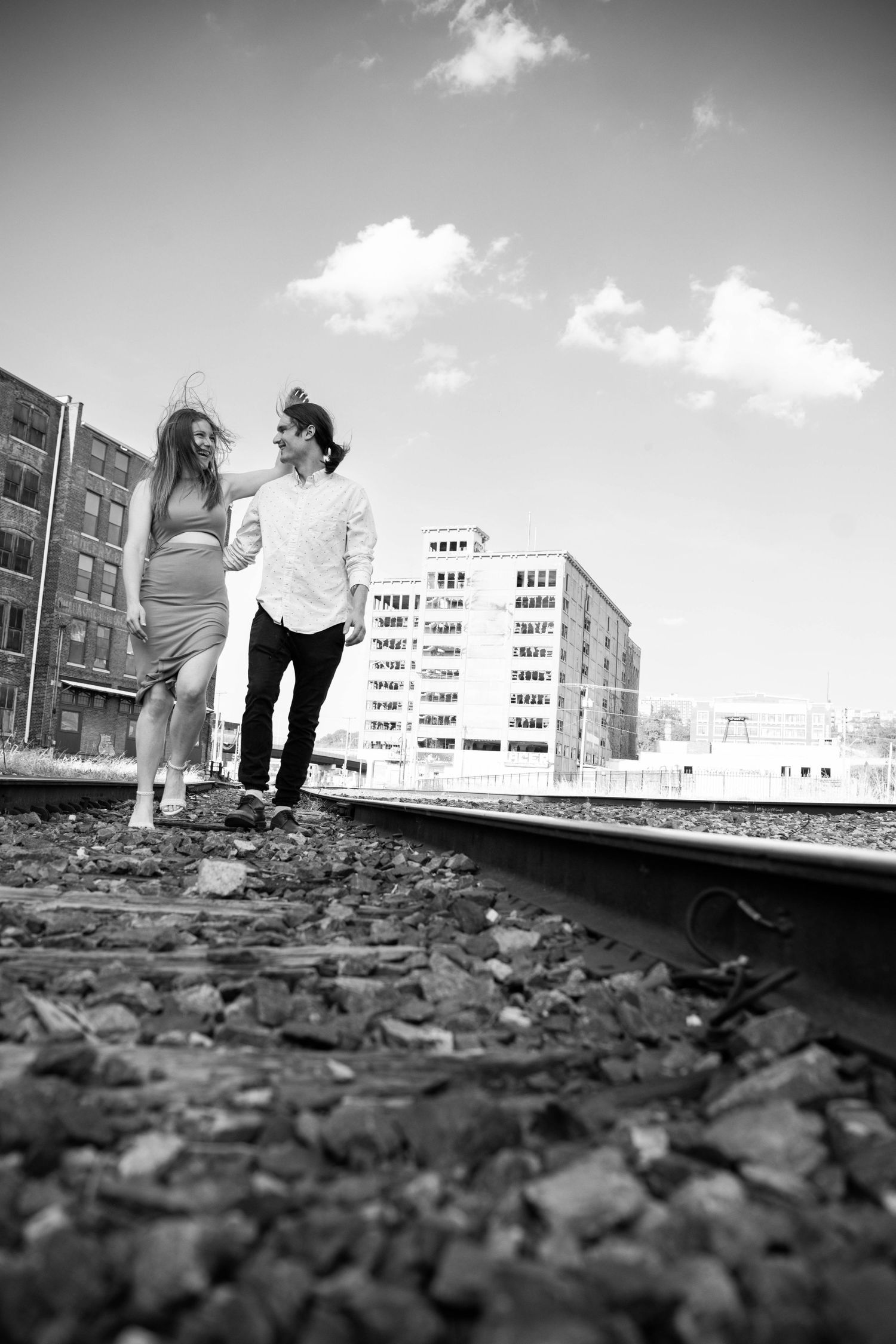 Black and white photo of a couple walking along railroad tracks with city buildings in background.