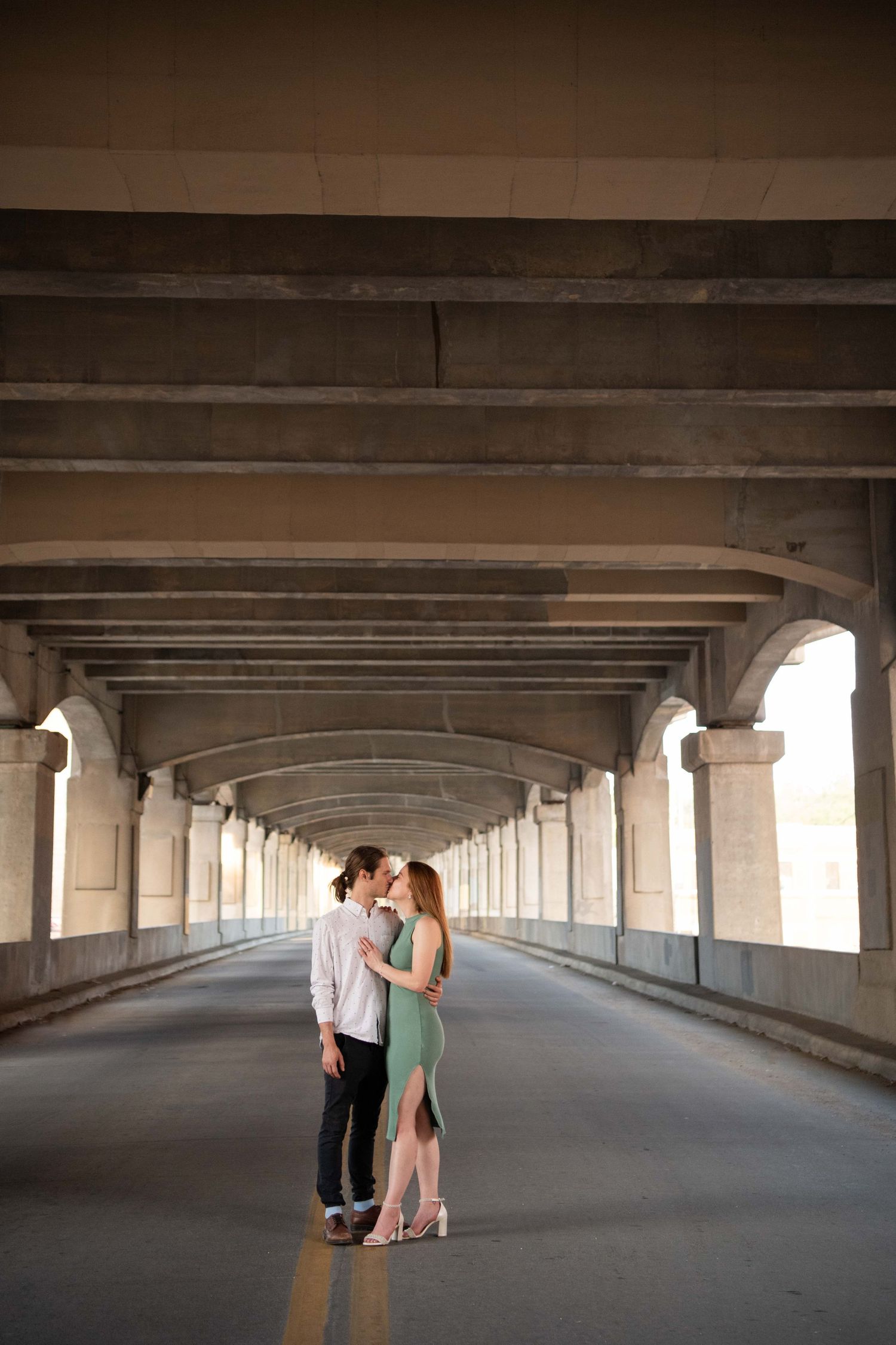 A couple stands in a long covered walkway with dramatic architectural columns and arches.