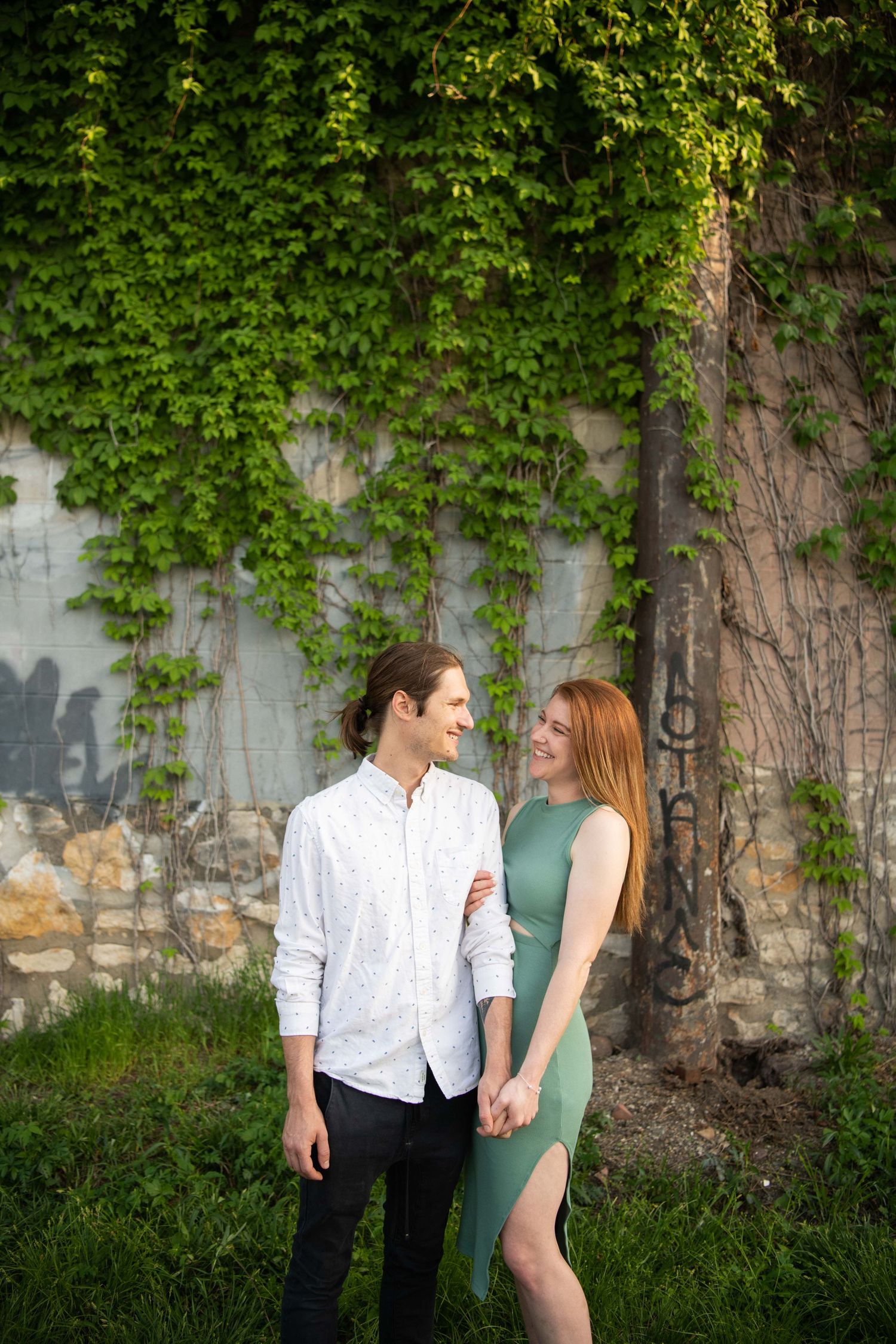A couple stands close together against an ivy-covered stone wall.