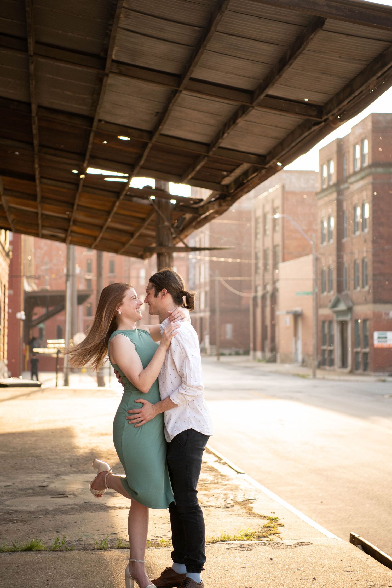 A romantic couple embraces under a rustic overhang as sunlight streams through in an urban setting.