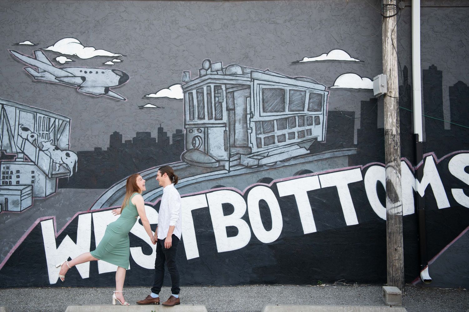 A couple poses playfully in front of an urban street art mural reading 'West Bottoms'.