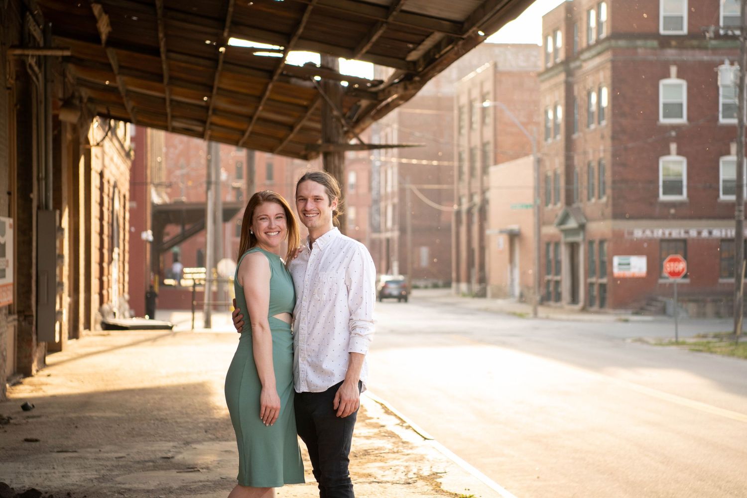 A couple poses together under an industrial overhang with brick buildings in the background.