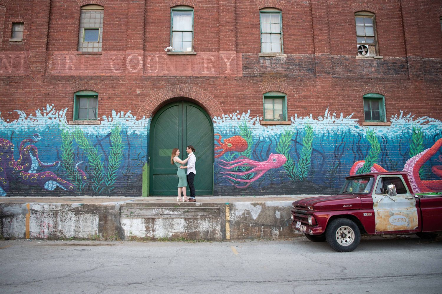 Couple stands on platform against brick wall with ocean mural and vintage truck in foreground.