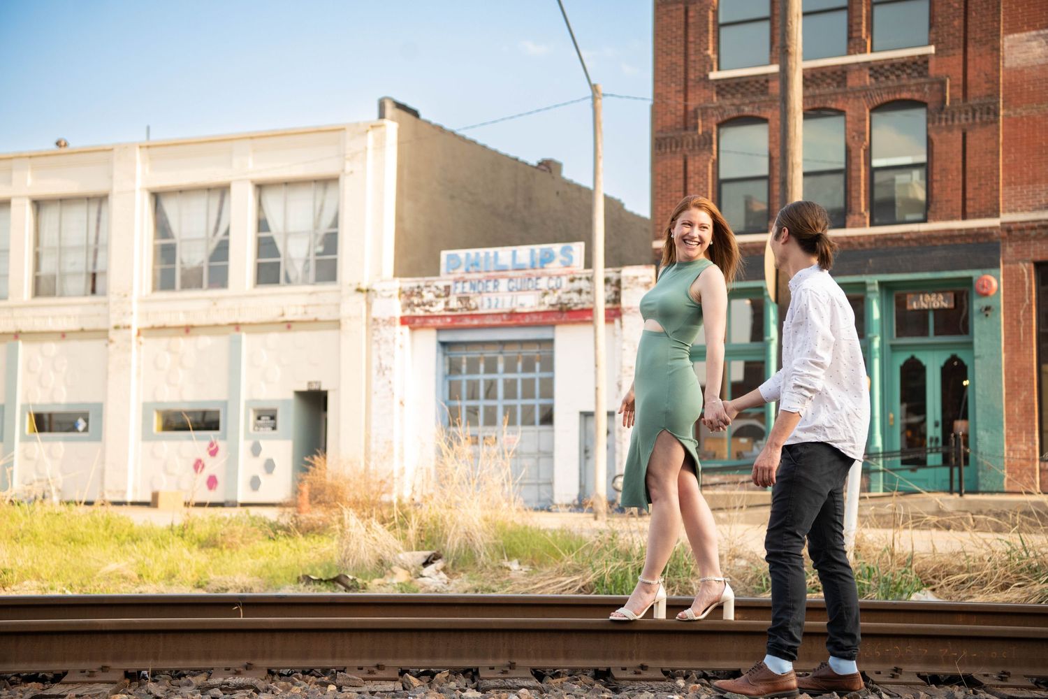 Couple poses together near railroad tracks with vintage storefronts and buildings in background.