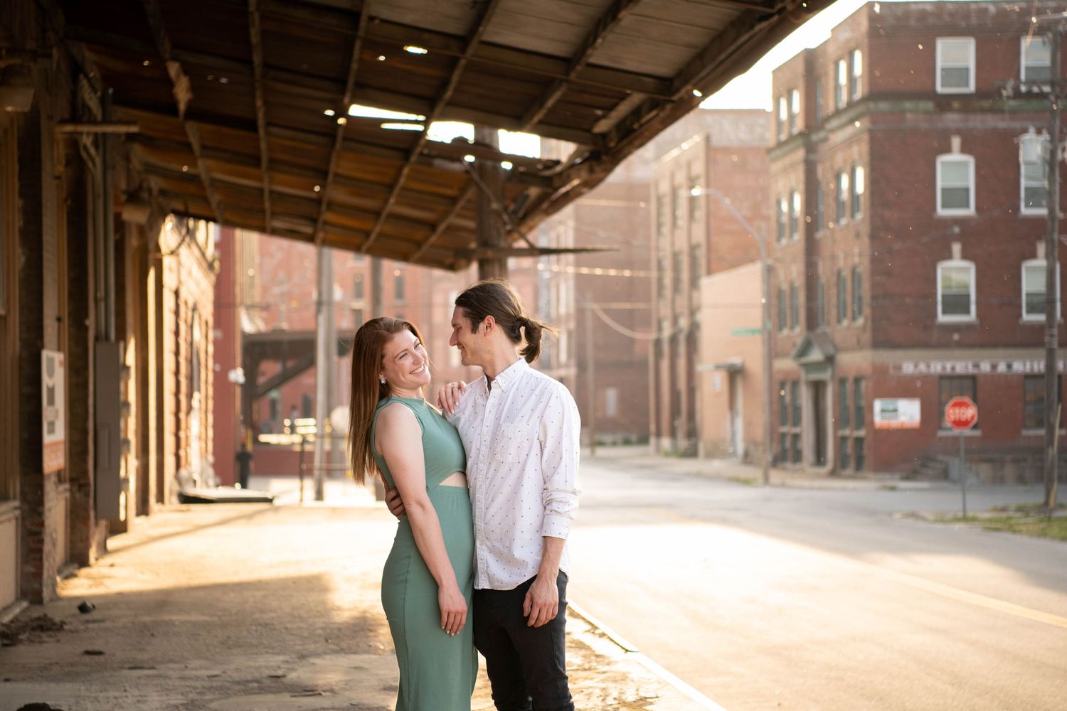 A couple shares a tender moment in an industrial setting with brick buildings in the background.