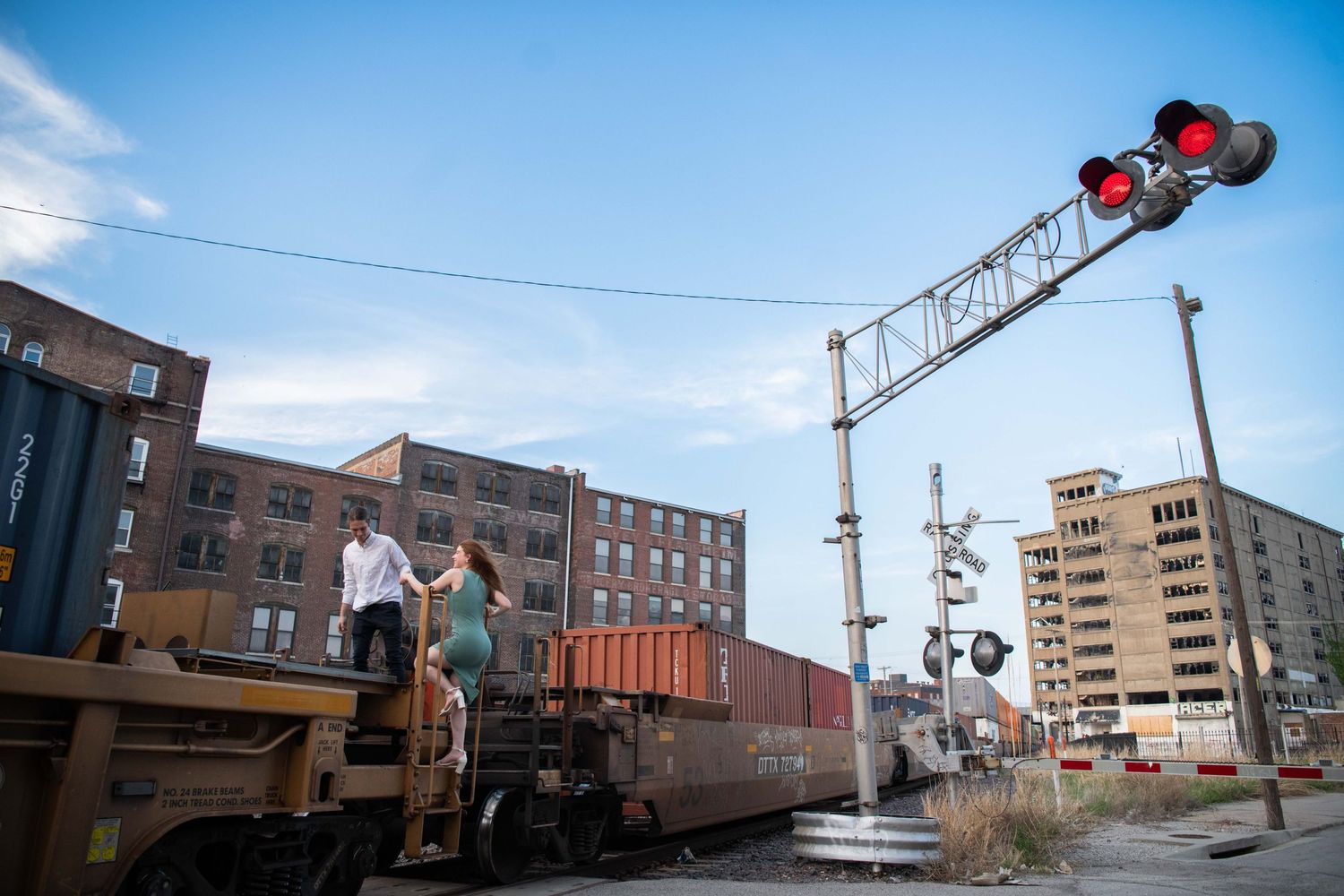 Couple standing on train car with railroad crossing signal and urban buildings in background.
