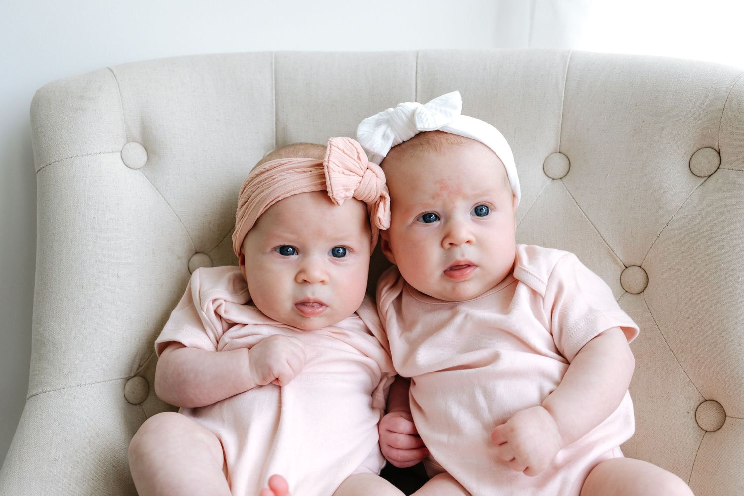 Two babies in matching pink outfits and bows sitting on a tufted cream chair.