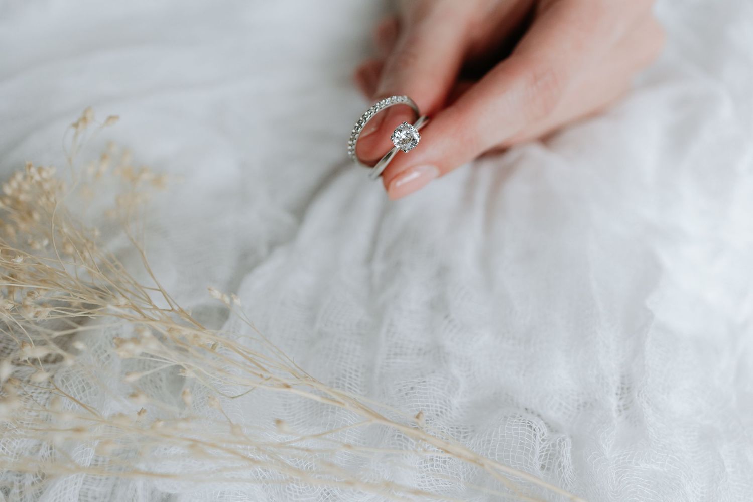 Close-up of a delicate ring being held against white textured fabric with dried flowers.
