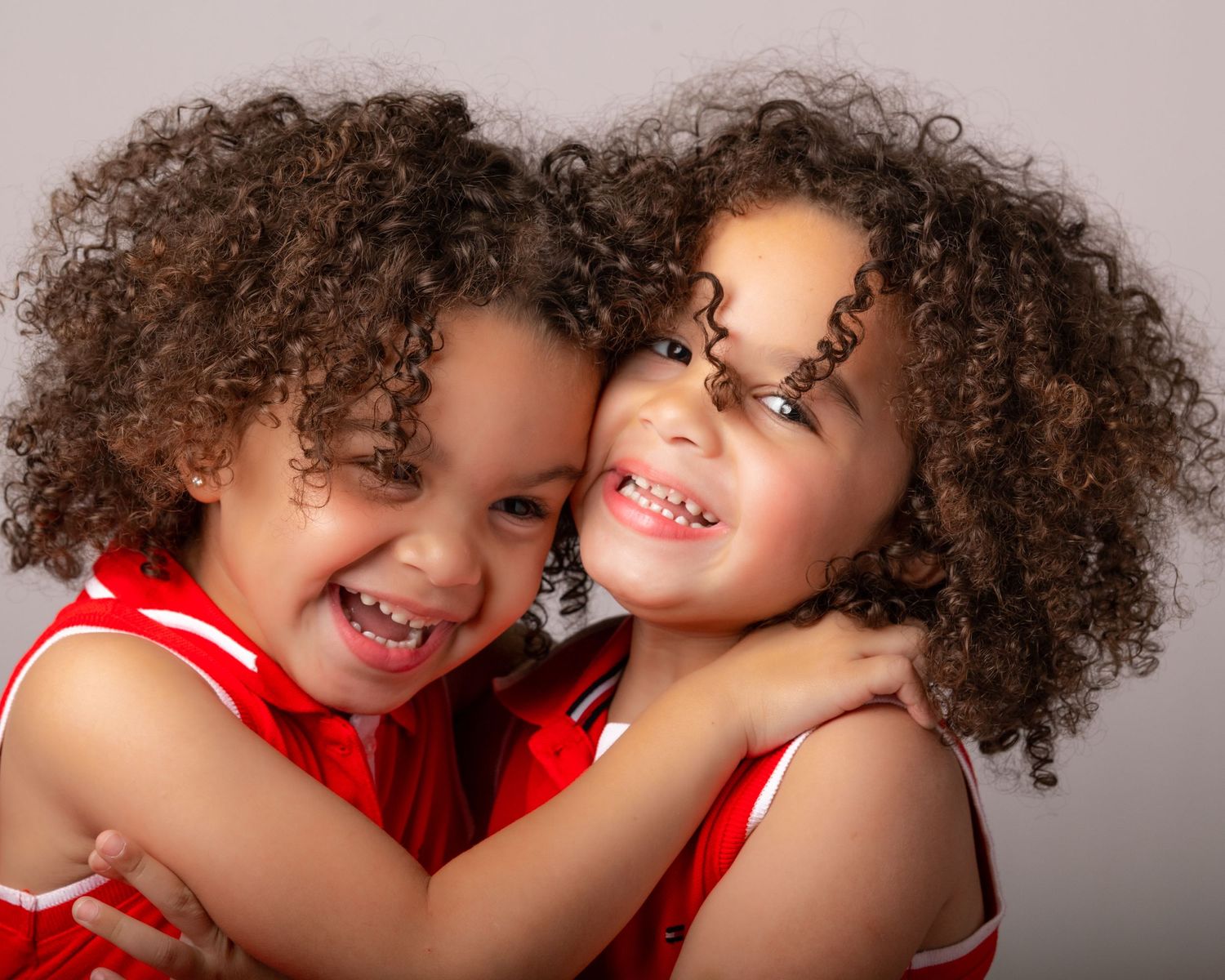 Two children with curly hair wearing red outfits hugging and smiling.