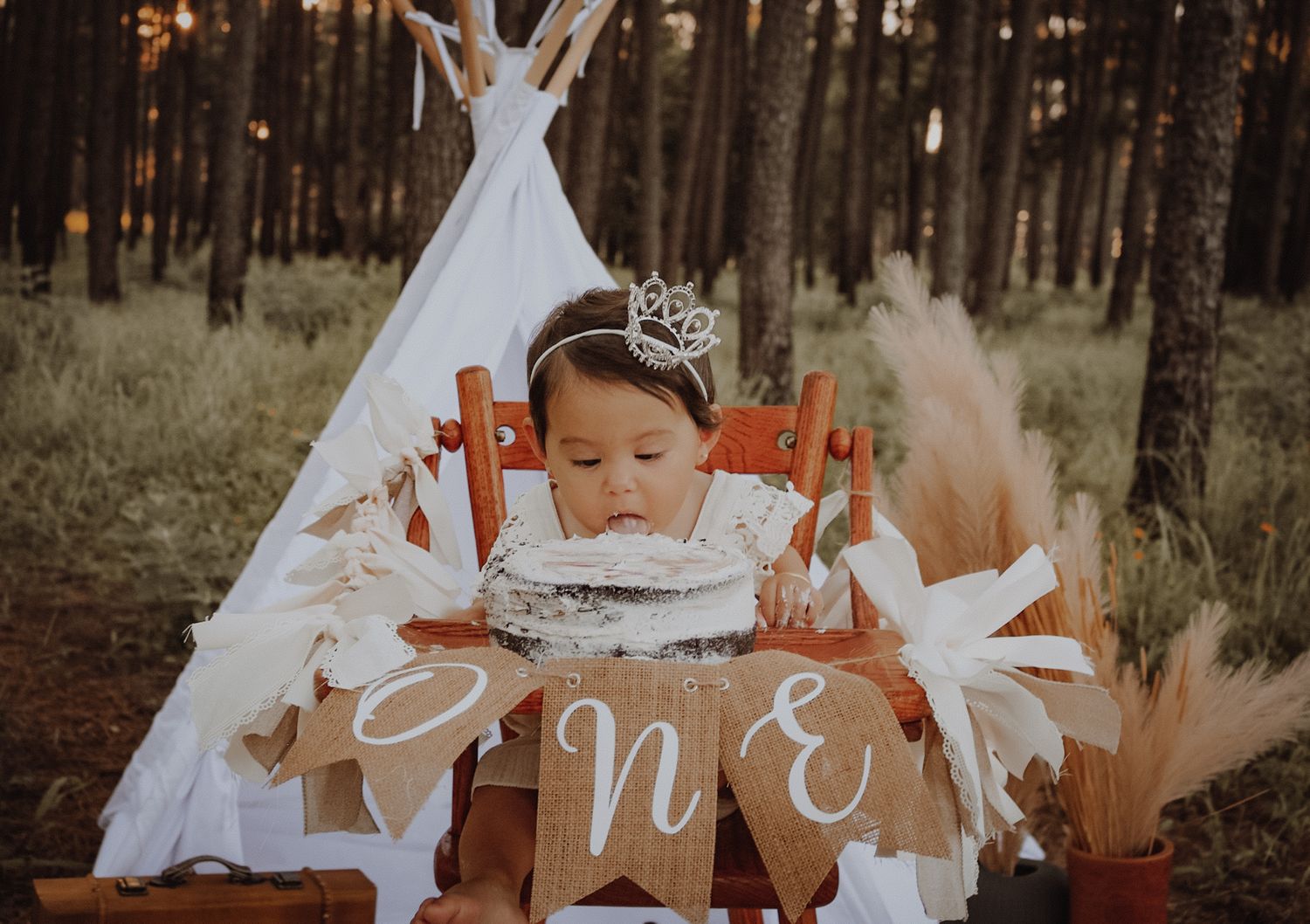 A teepee tent setup in a forest with 'ONE' banner and boho decorations for a first birthday celebration.