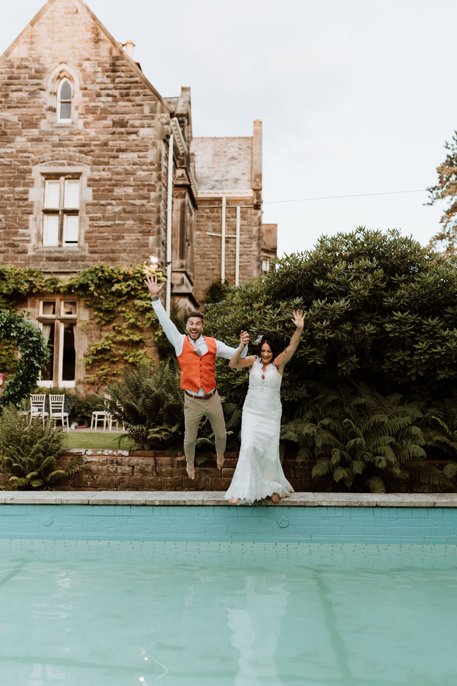 A cool couple jumps with arms raised by a swimming pool in front of a stone mansion on their wedding day at Dunwood Hall