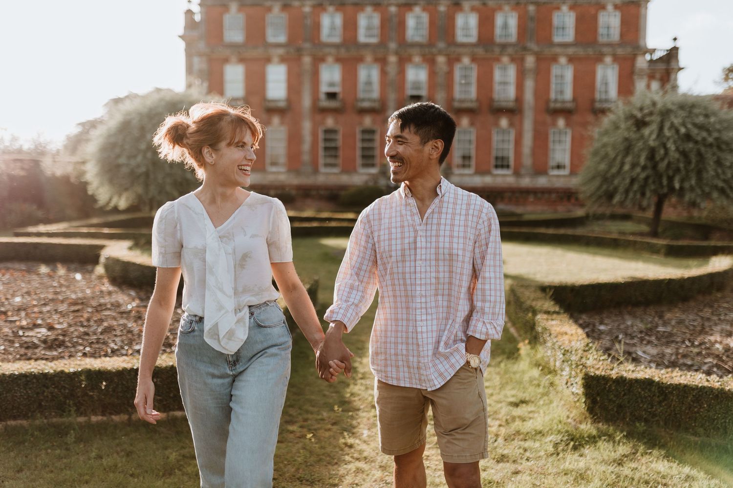 A couple holds hands while walking together through a garden with Berwick House in the background on a sunny day
