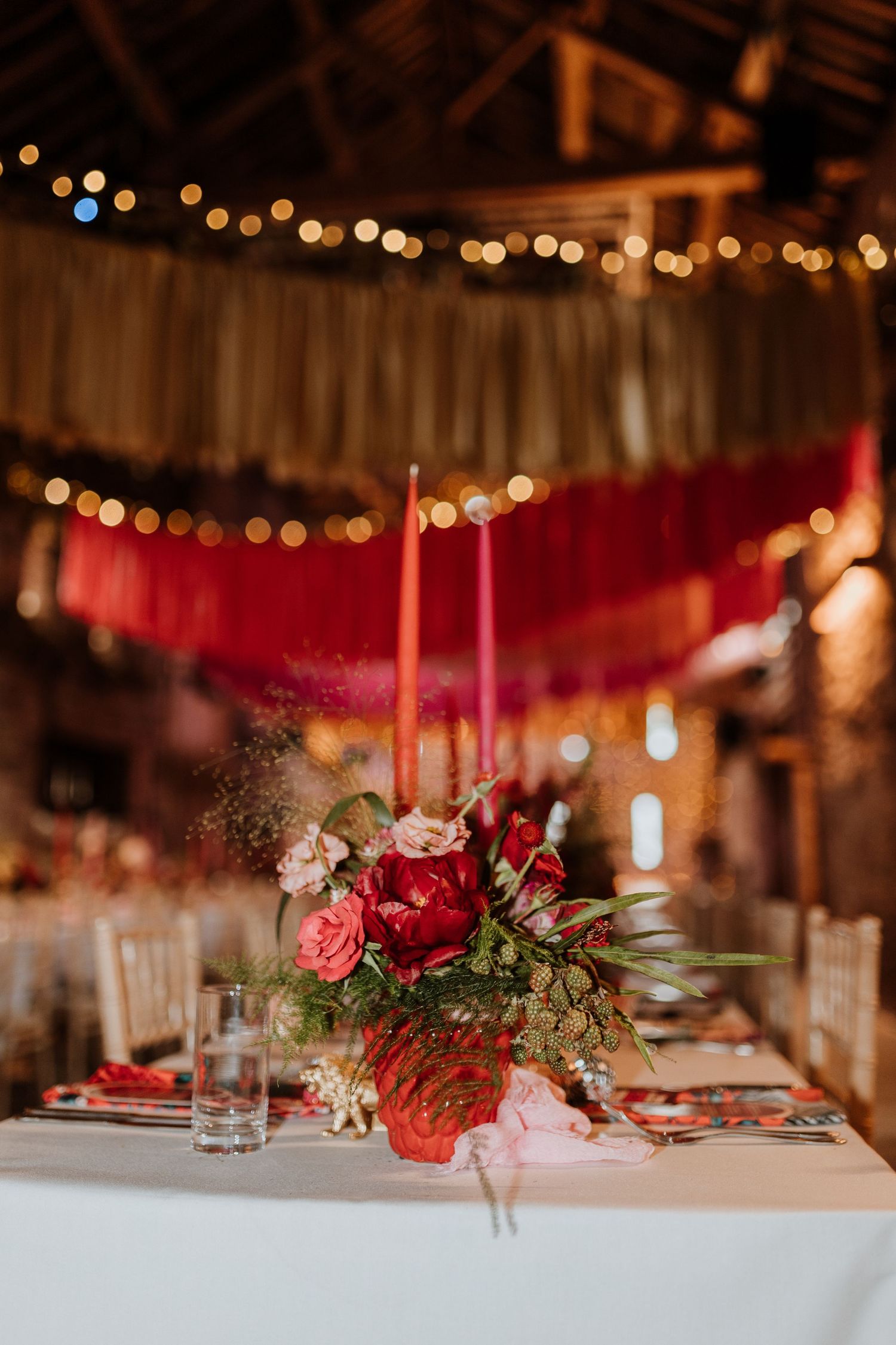 Red floral centerpiece with candles on white tablecloth at wedding reception with string lights overhead in Lyde Court