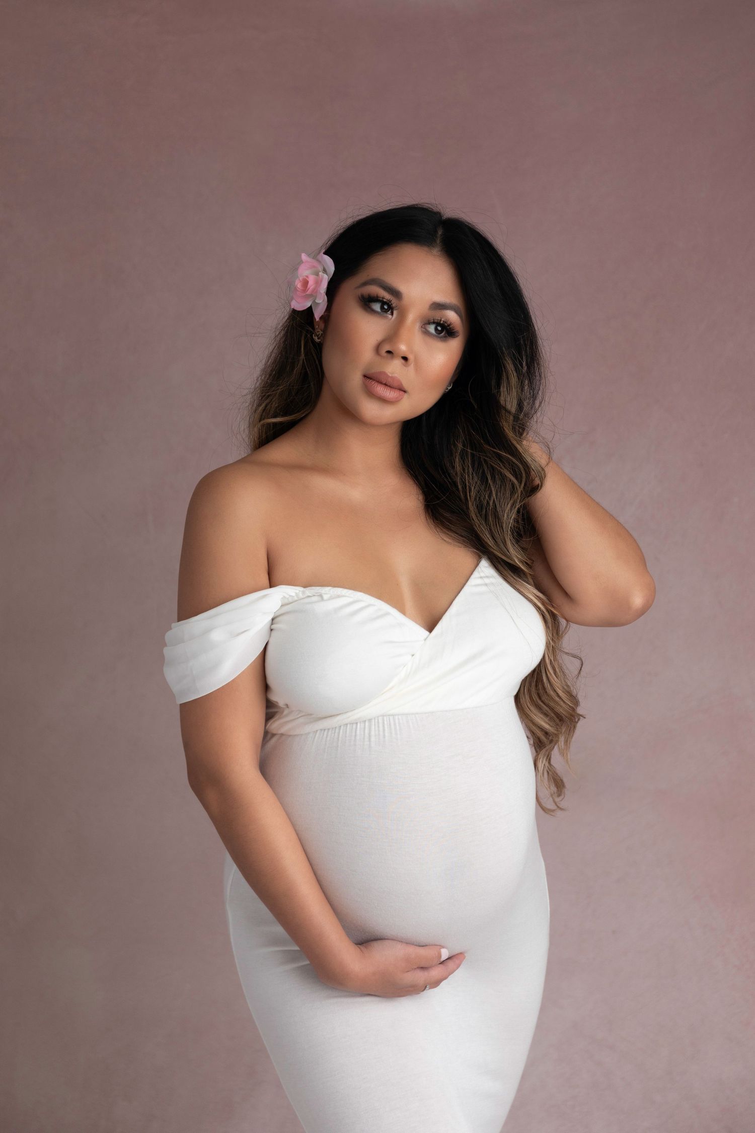 Maternity portrait of woman in white off-shoulder dress against mauve backdrop.