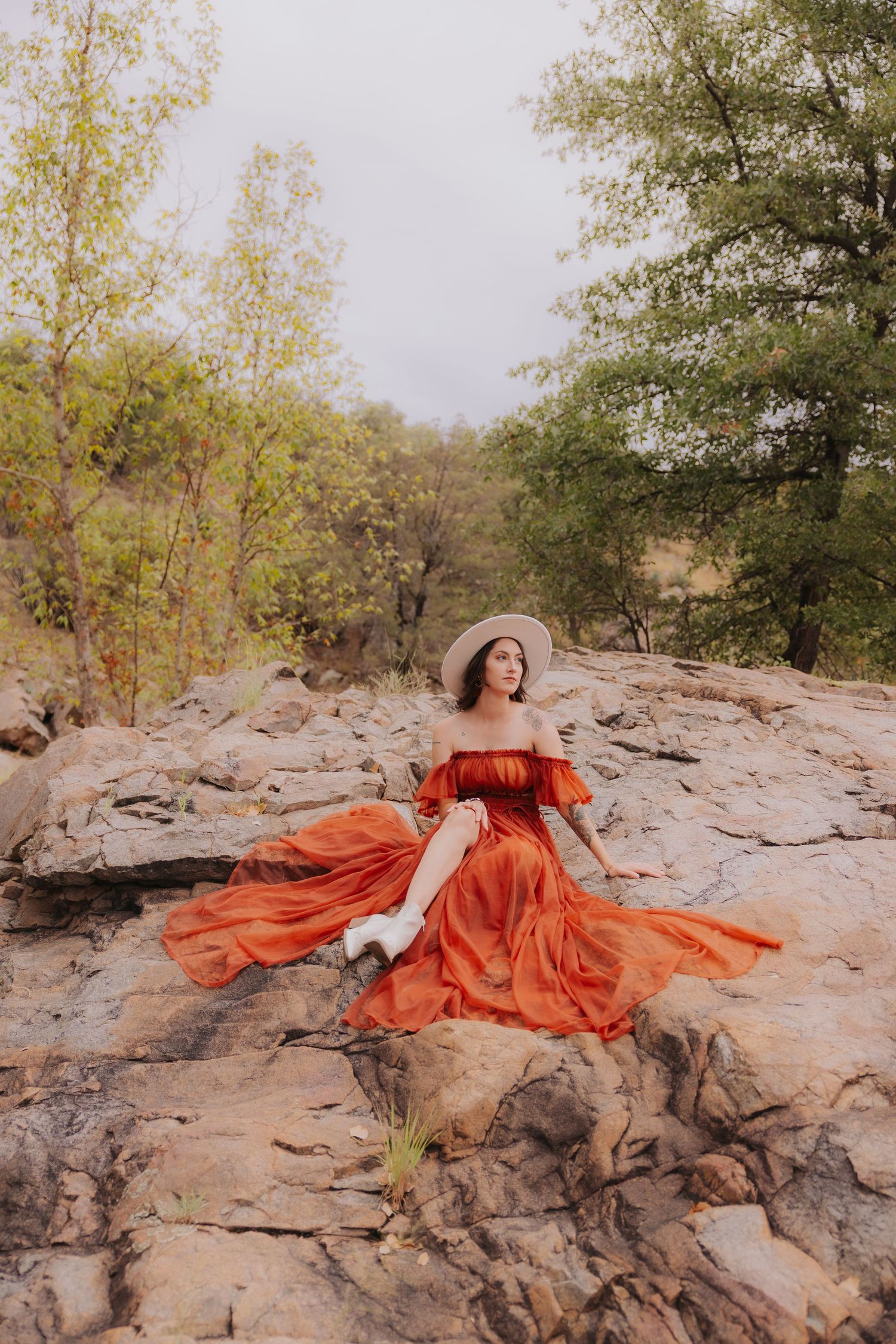 Person in flowing orange dress poses dramatically on rocky terrain surrounded by trees.