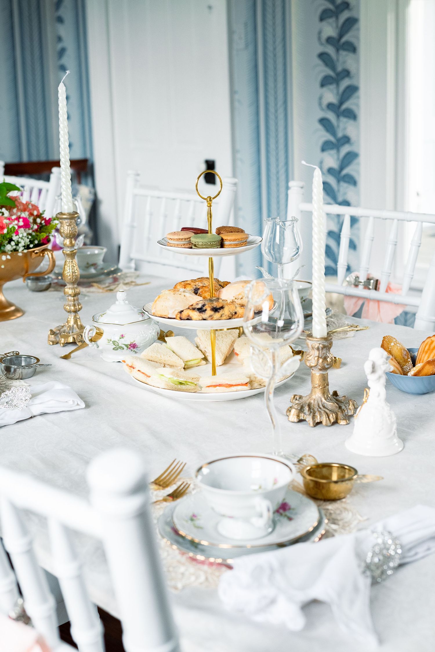 Tiered serving tray with sandwiches and treats on an elegant white tablecloth with gold accents.
