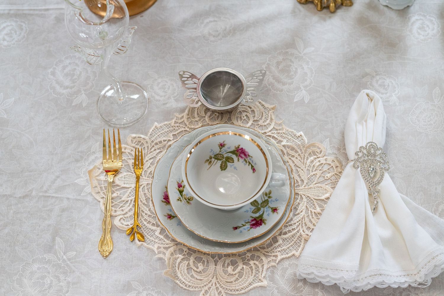 Vintage place setting with floral teacup, gold cutlery, and white napkin on lace charger.